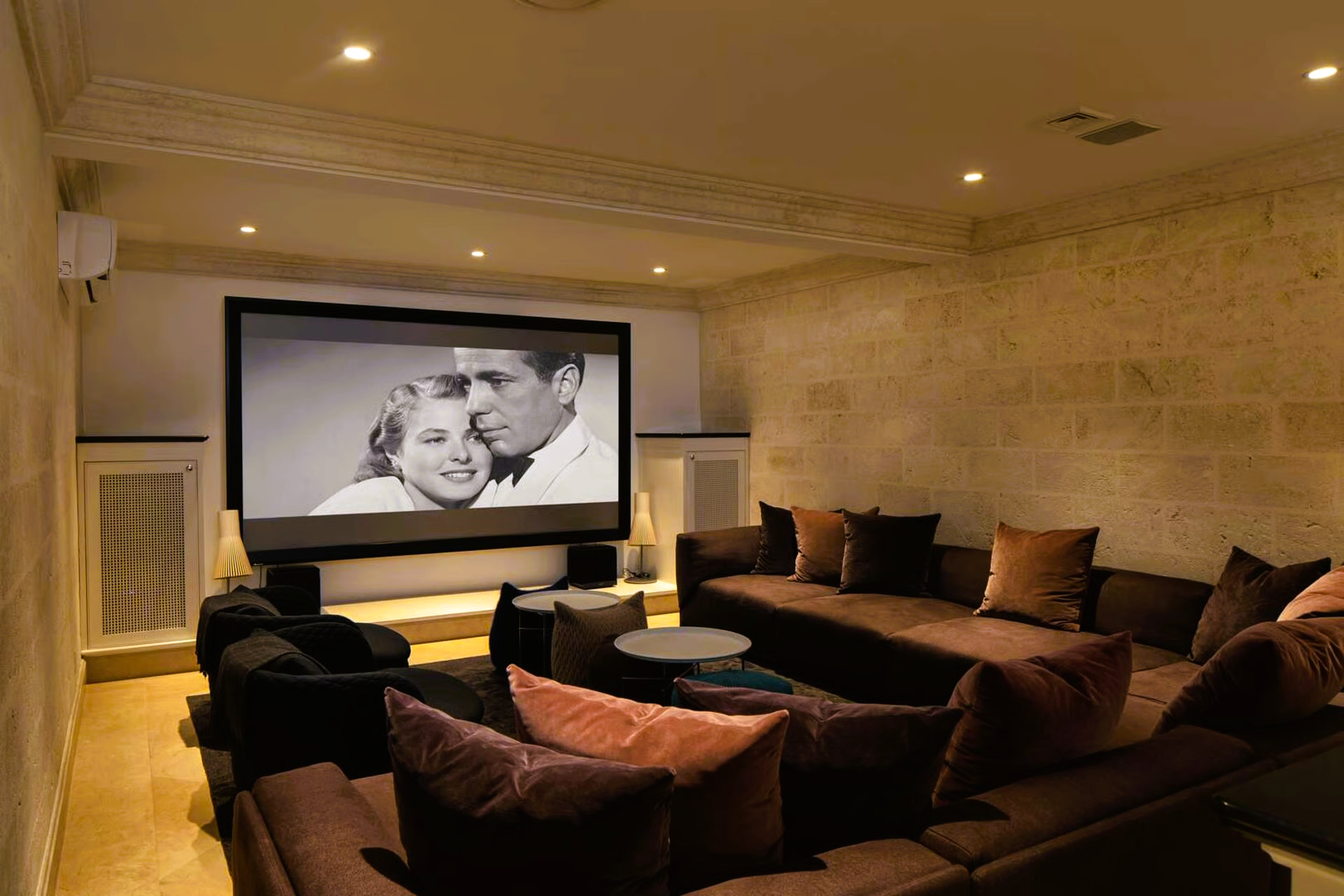 Home theater with large screen showing a black and white movie, brown sectional seating, and stone walls.