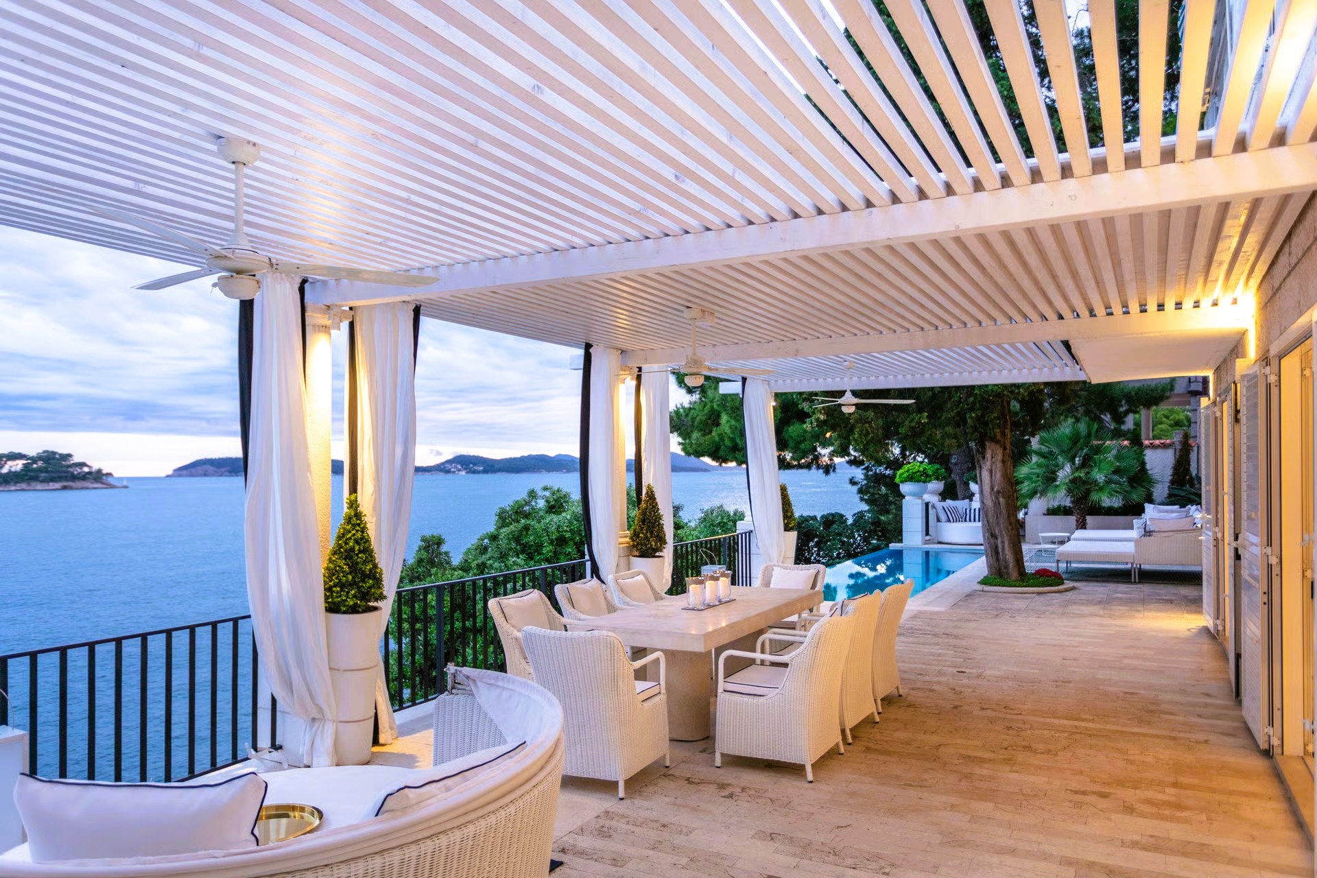 Outdoor dining area overlooking the sea, with white furniture, curtains, and a swimming pool.