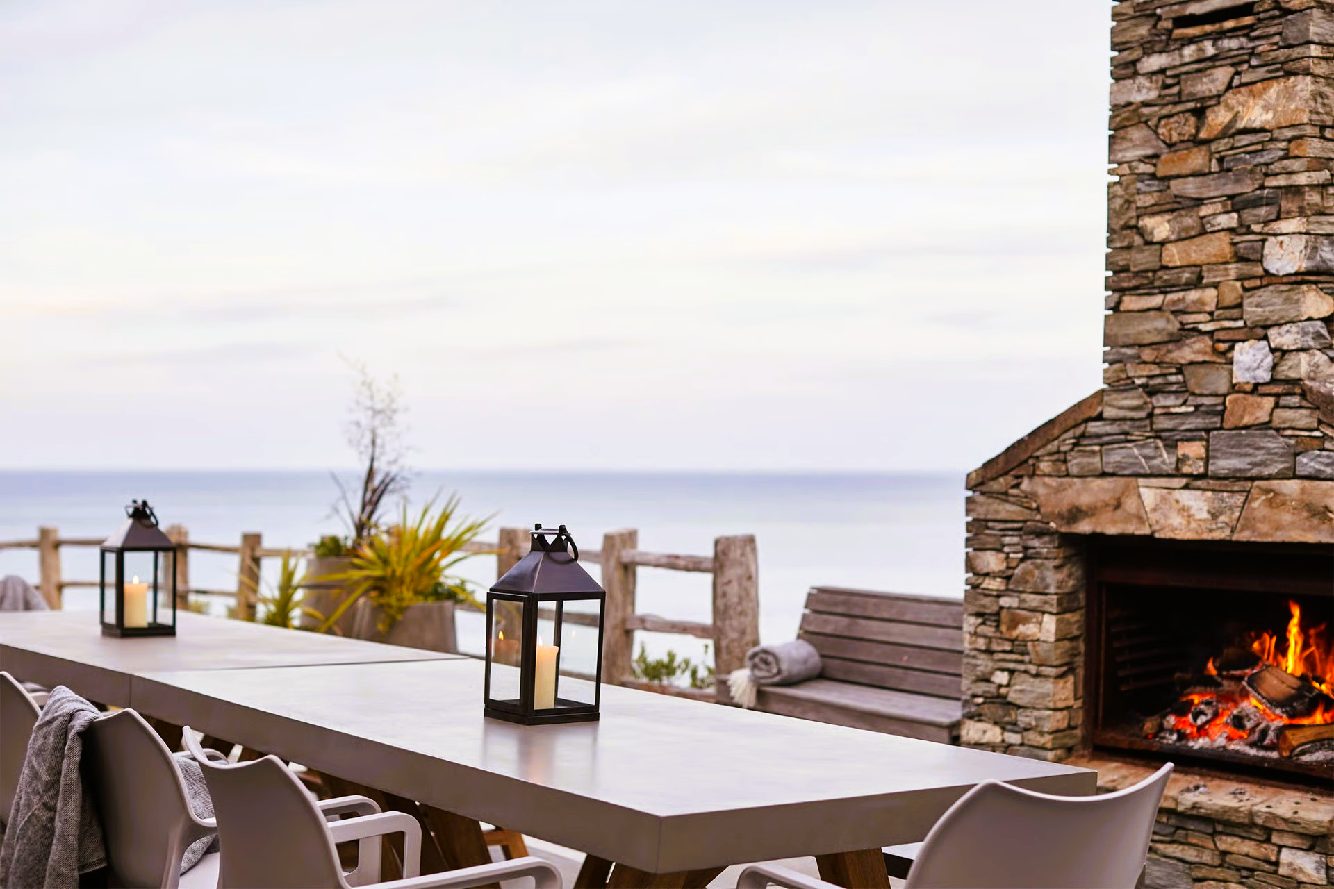 Outdoor dining area with ocean view and stone fireplace, lanterns on table.