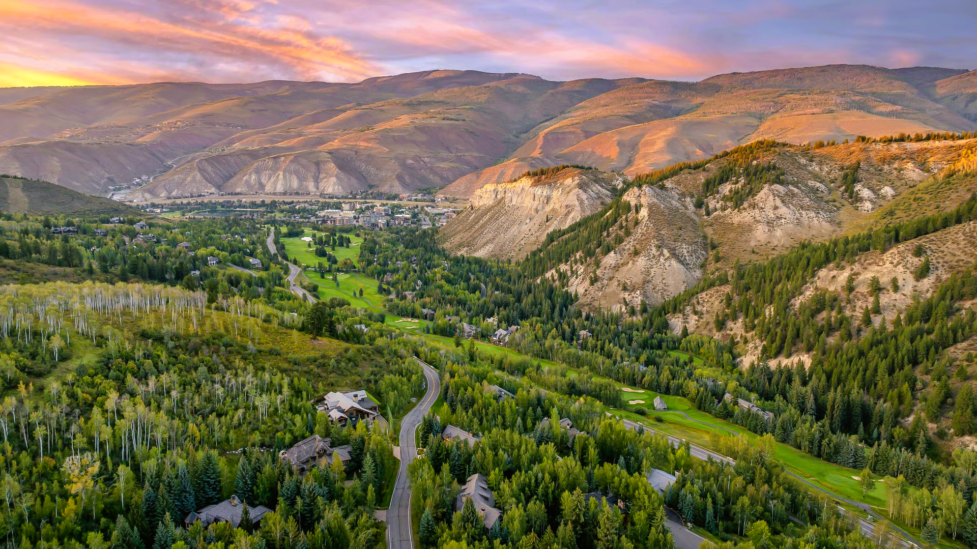 Green valley with a road and golf course, surrounded by mountains at sunset.