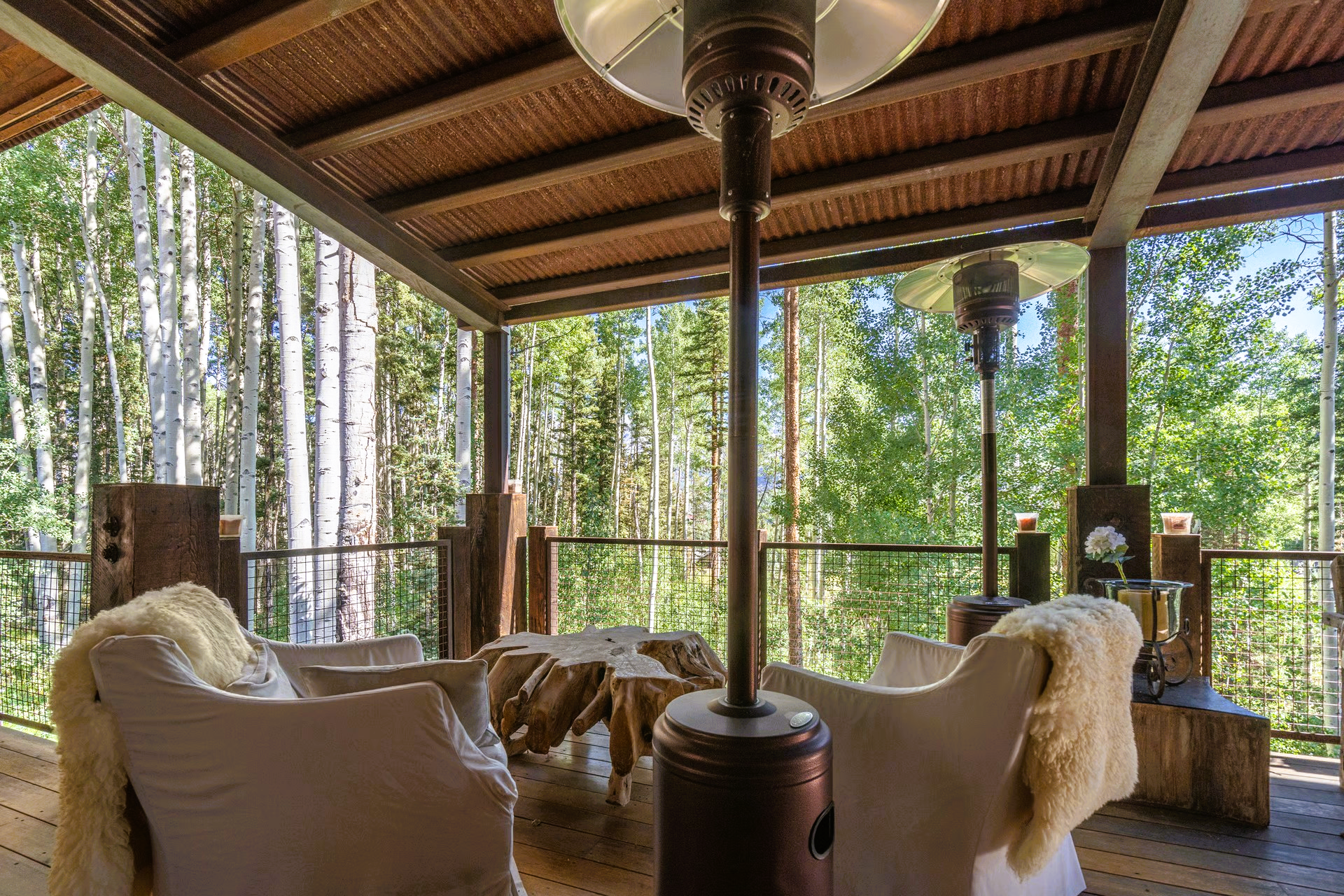 Outdoor seating area with a view of trees. Cozy white chairs, a wood table, and patio heaters.