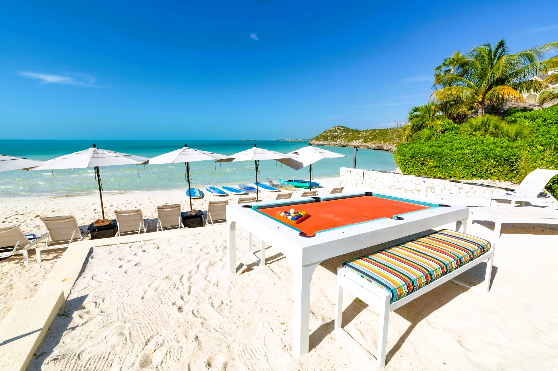 Pool table on a white sandy beach with umbrellas, lounge chairs, and clear turquoise water under a blue sky.