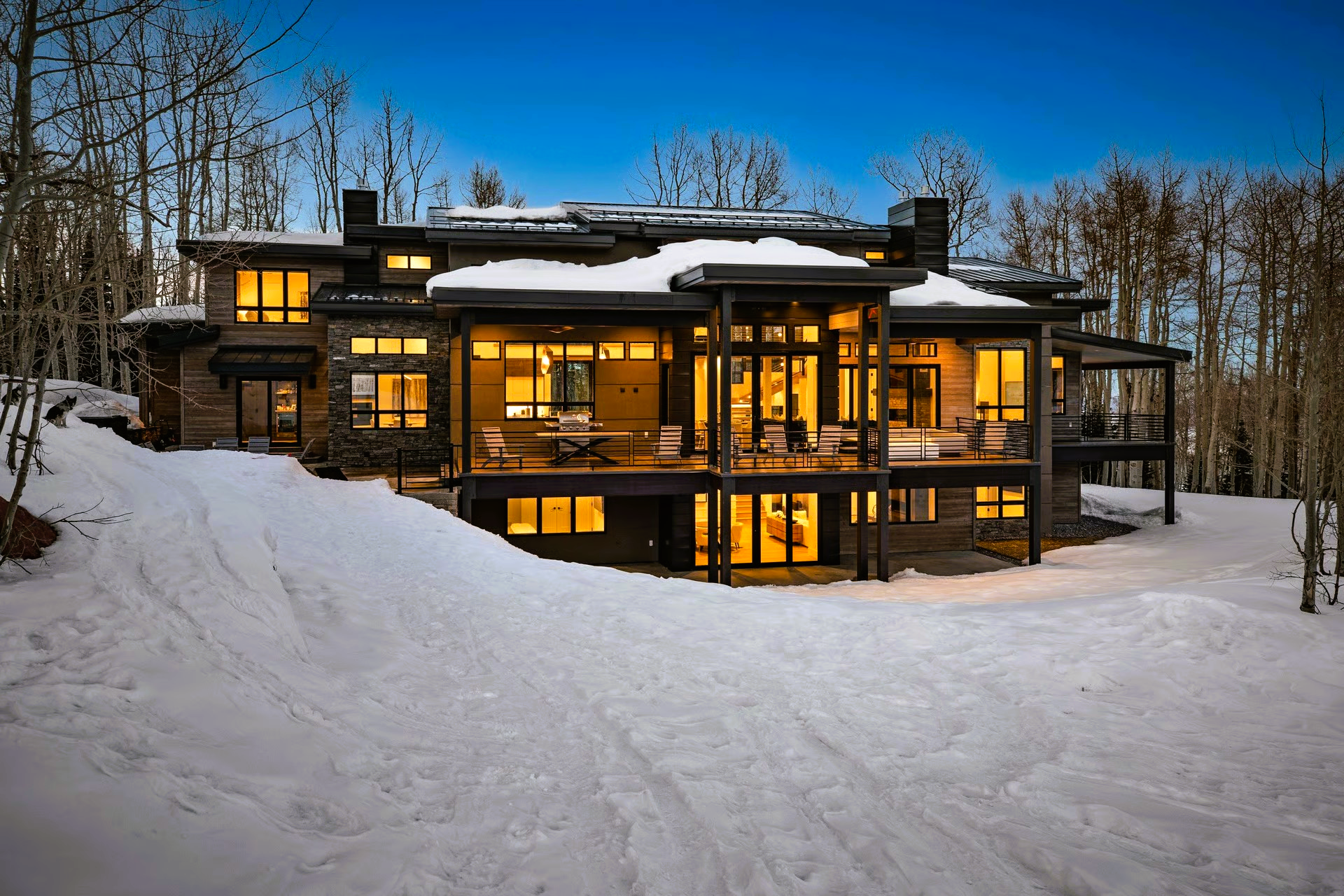 Snowy house at dusk with lit windows, surrounded by trees.