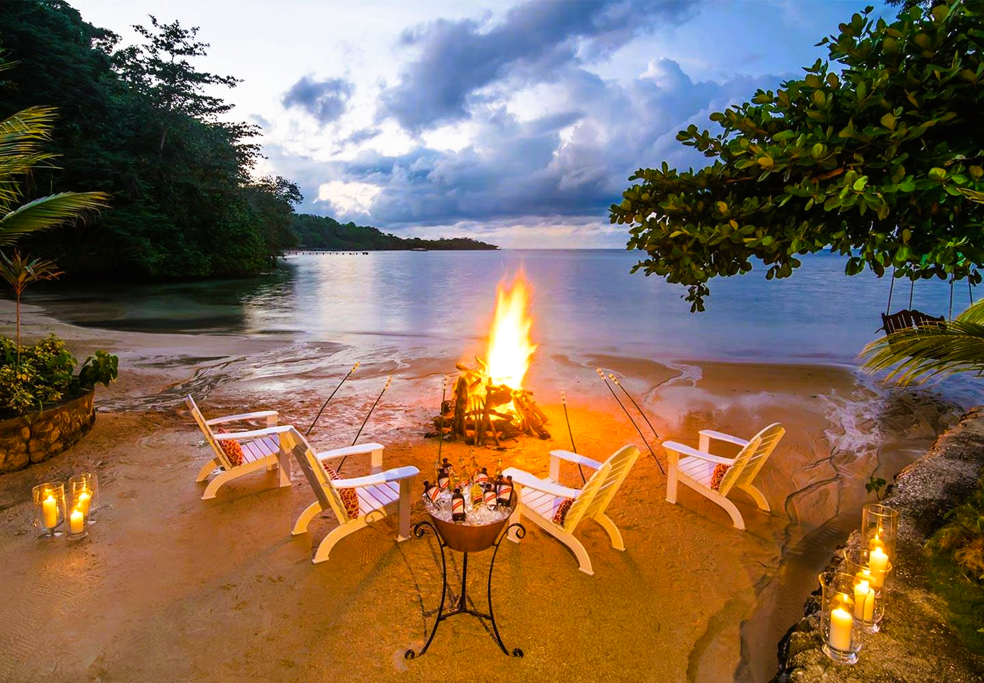 Beach scene: fire pit, chairs, drinks, calm water, evening sky.