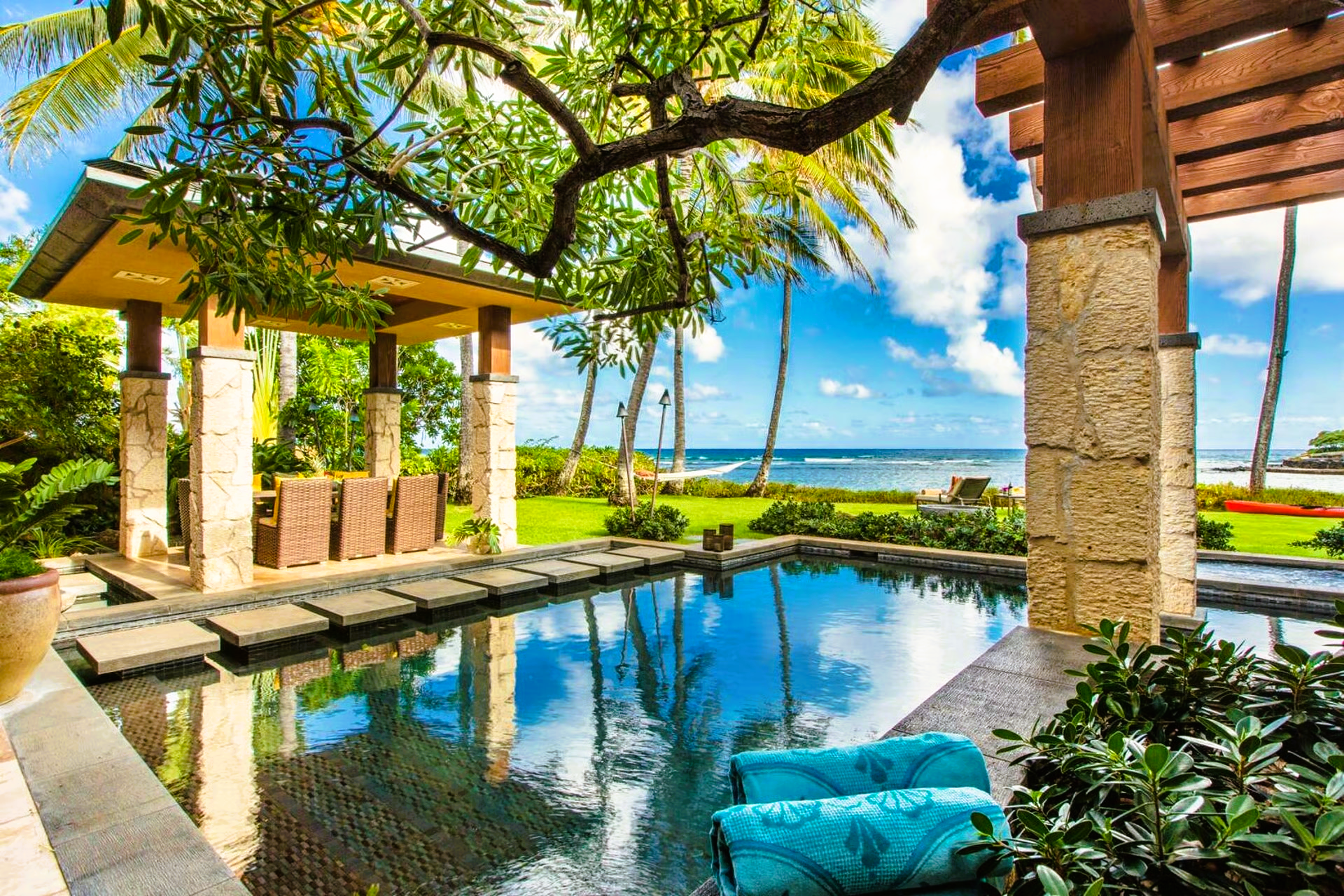 Poolside view: turquoise towels, gazebo, water reflecting blue sky and palm trees on a sunny day by the ocean.