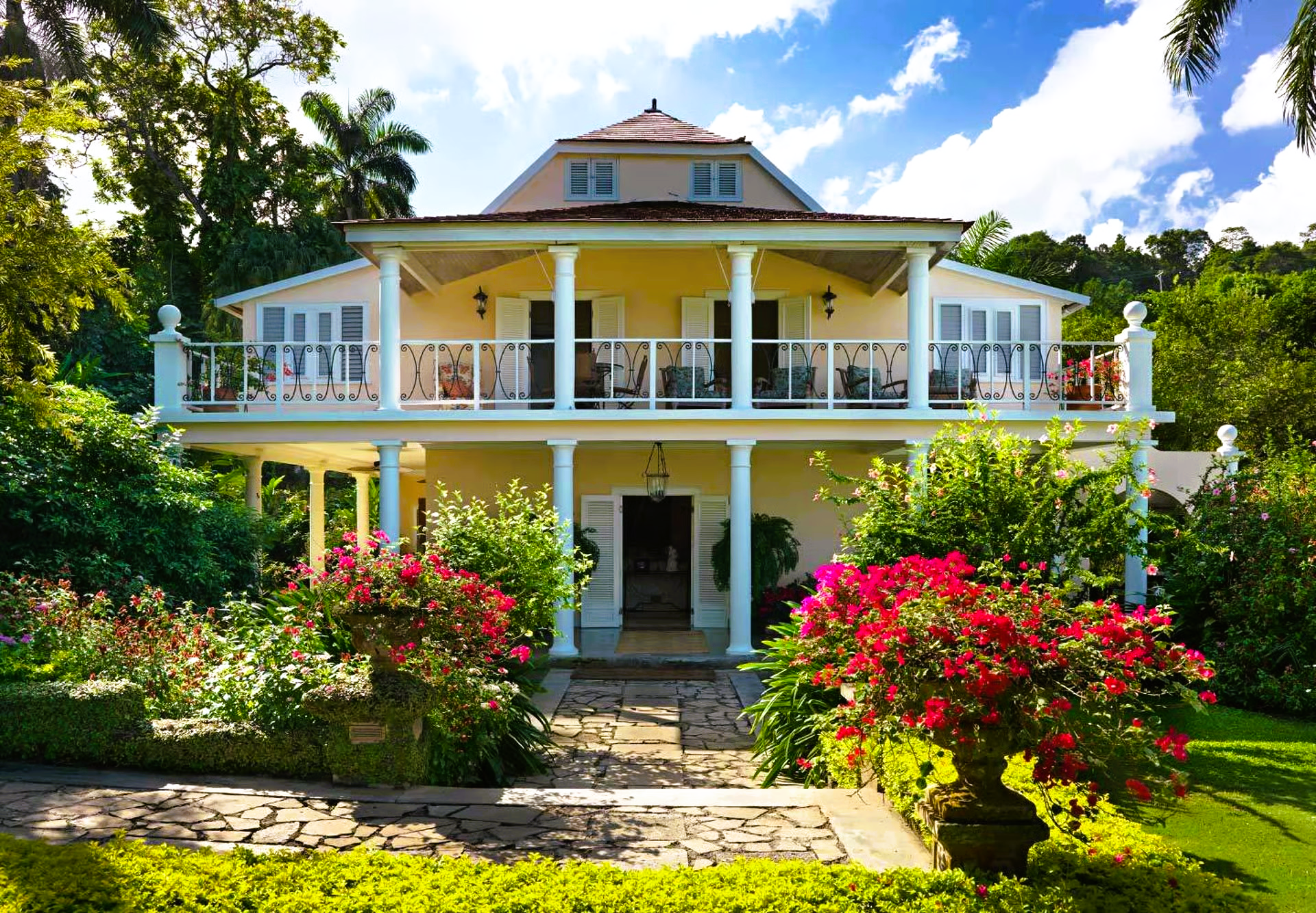Two-story beige house with columns, balcony, and colorful flower gardens under a blue sky.