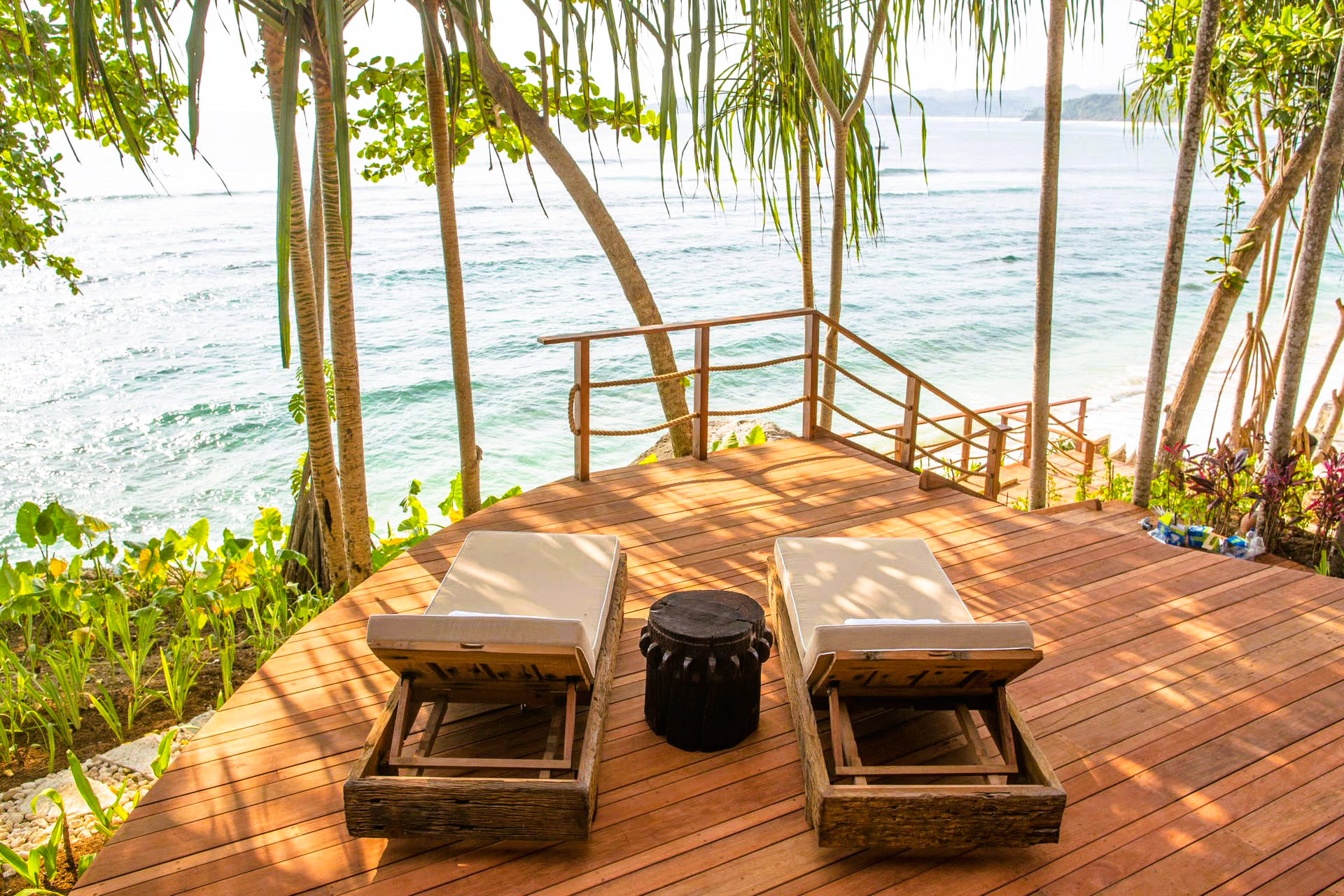 Two lounge chairs face the ocean on a wooden deck; water, trees and a small table are nearby.