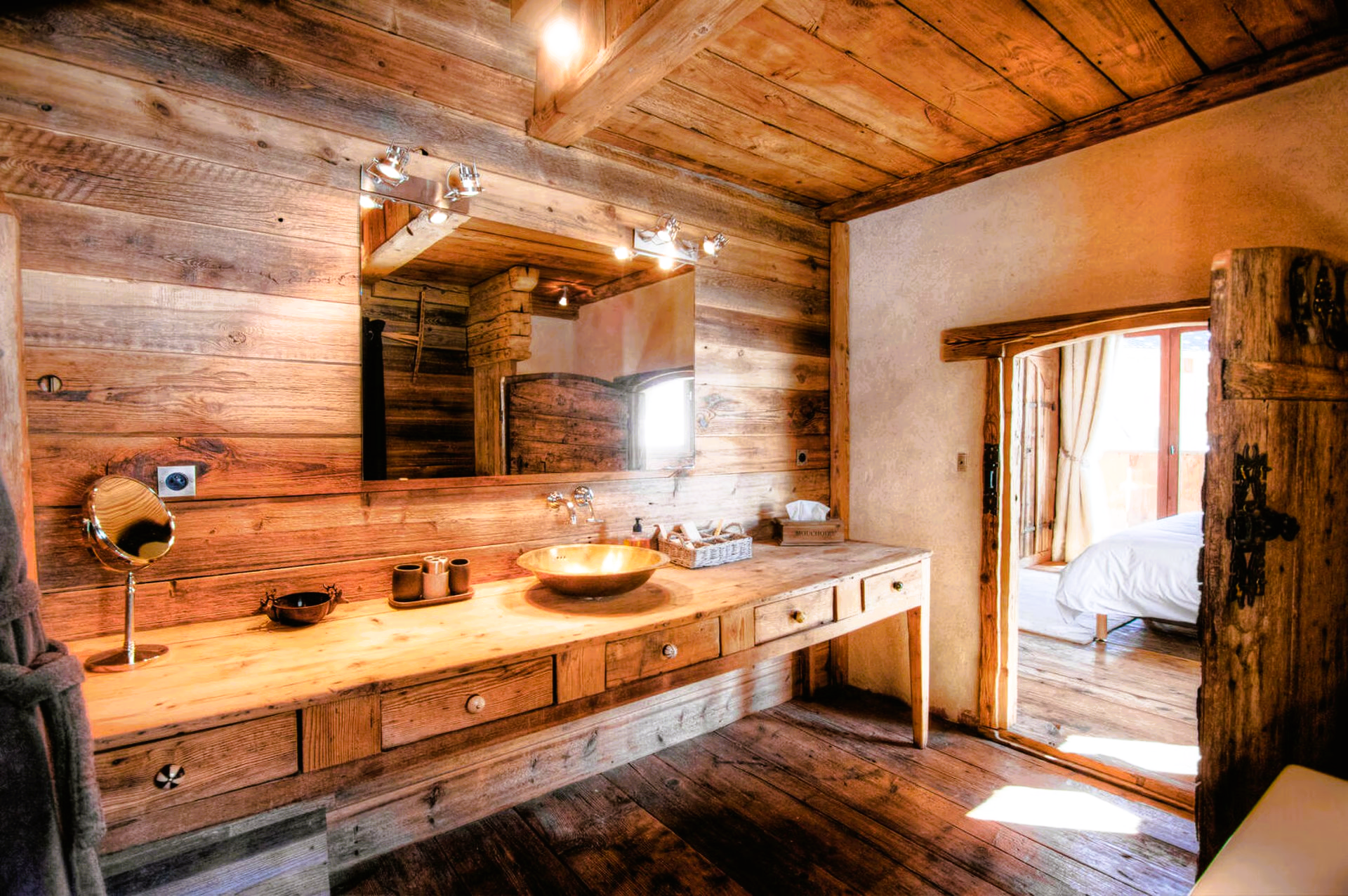 Rustic wooden bathroom with a long vanity, gold sink, and open doorway to a bedroom.