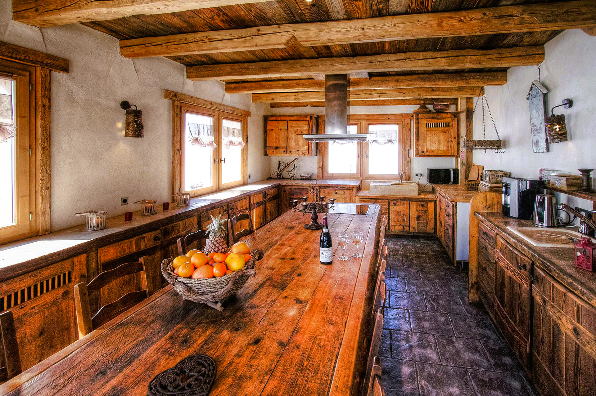 Rustic kitchen with long wooden table, cabinets, and exposed beams. Sunlight streams through windows.