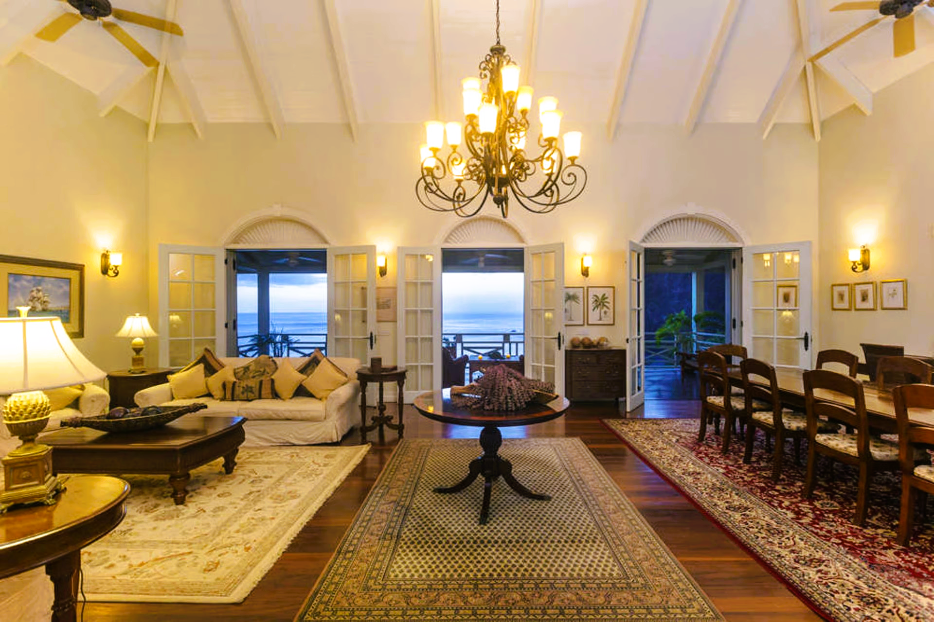 Elegant interior with ocean view: living and dining areas, beige walls, chandelier, wood floors, open doors.