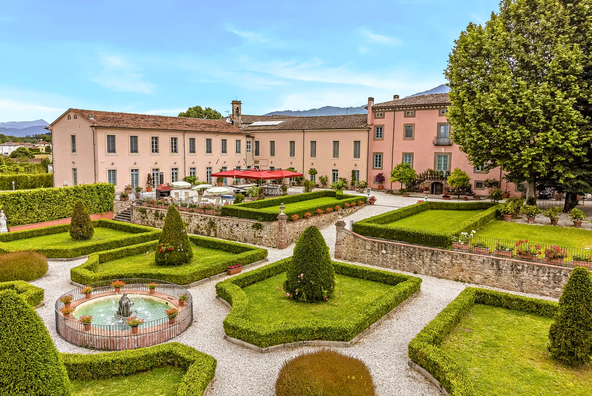Formal garden with hedges, a fountain, and a pink building in Italy.