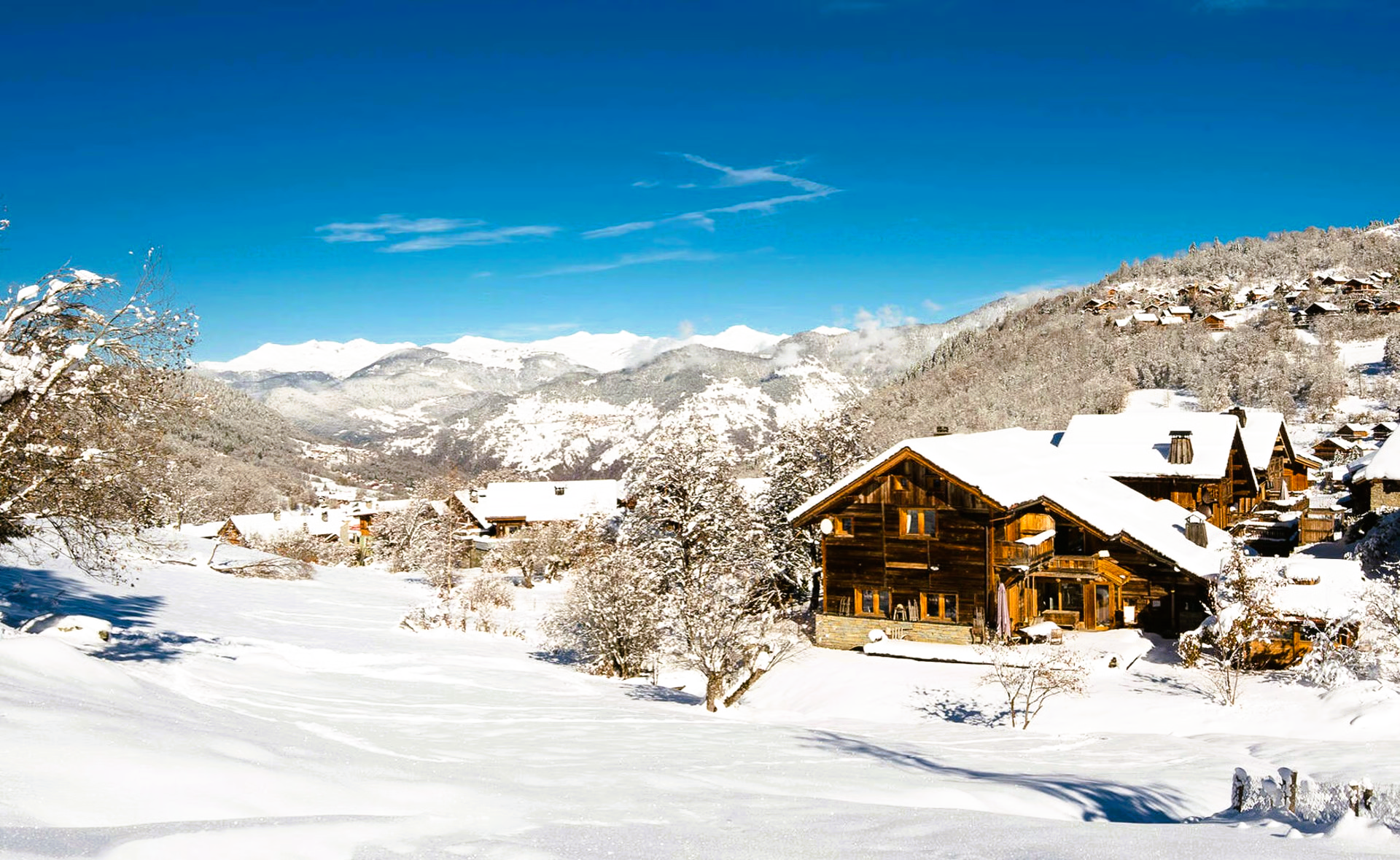 Snow-covered village nestled in a valley with wooden houses and mountain backdrop under a bright blue sky.