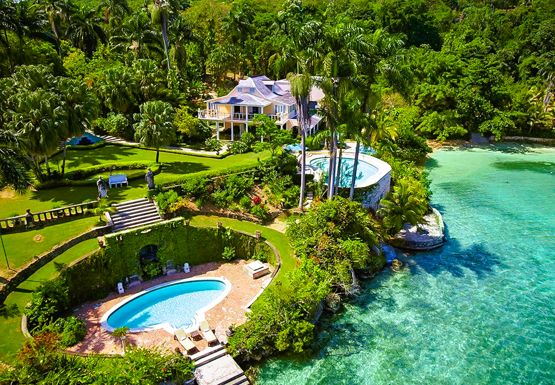Luxurious waterfront villa with two pools, lush greenery, and turquoise water.