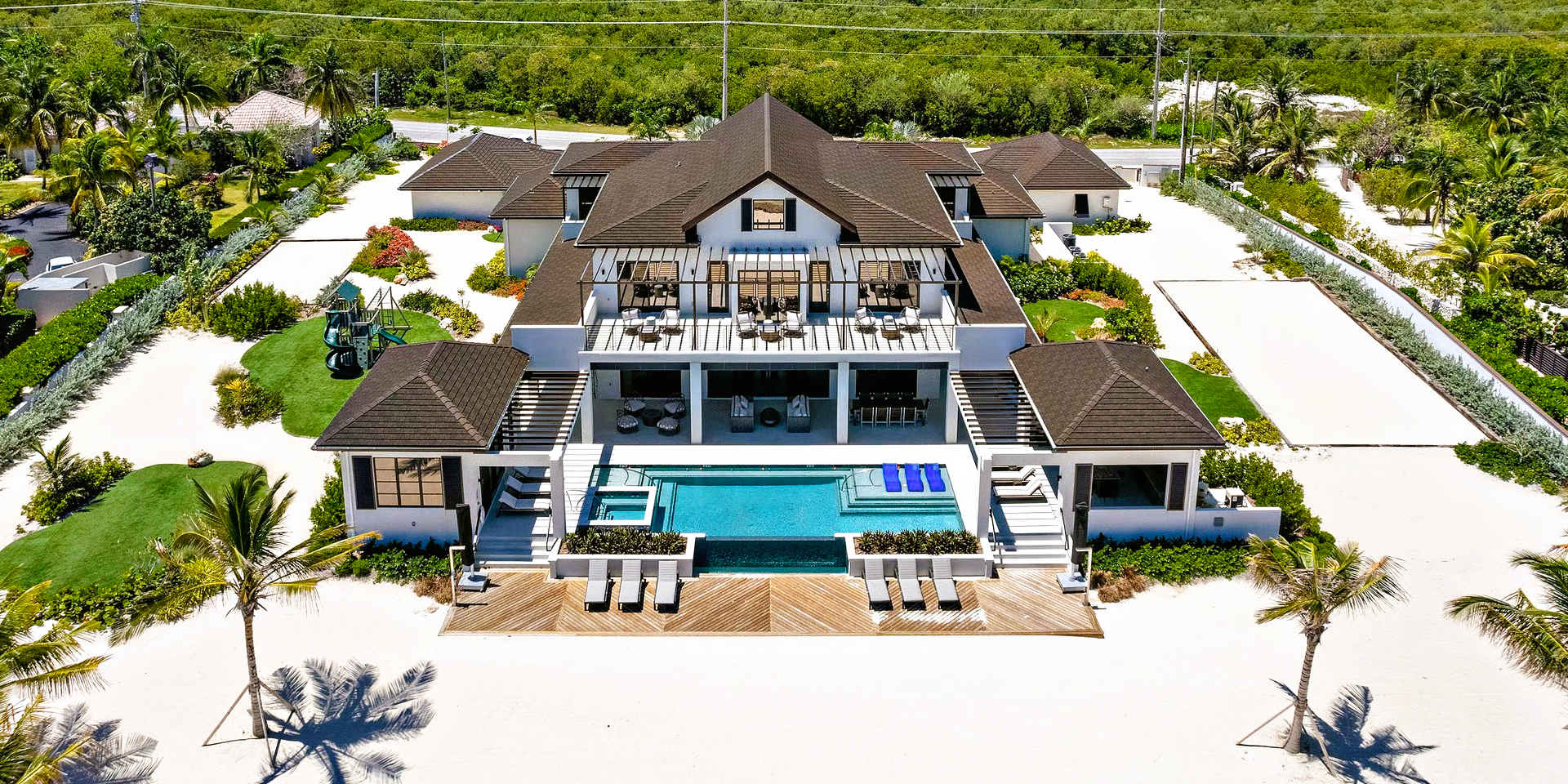 Aerial view of a luxurious white house with a pool and deck on a sandy beach with palm trees.