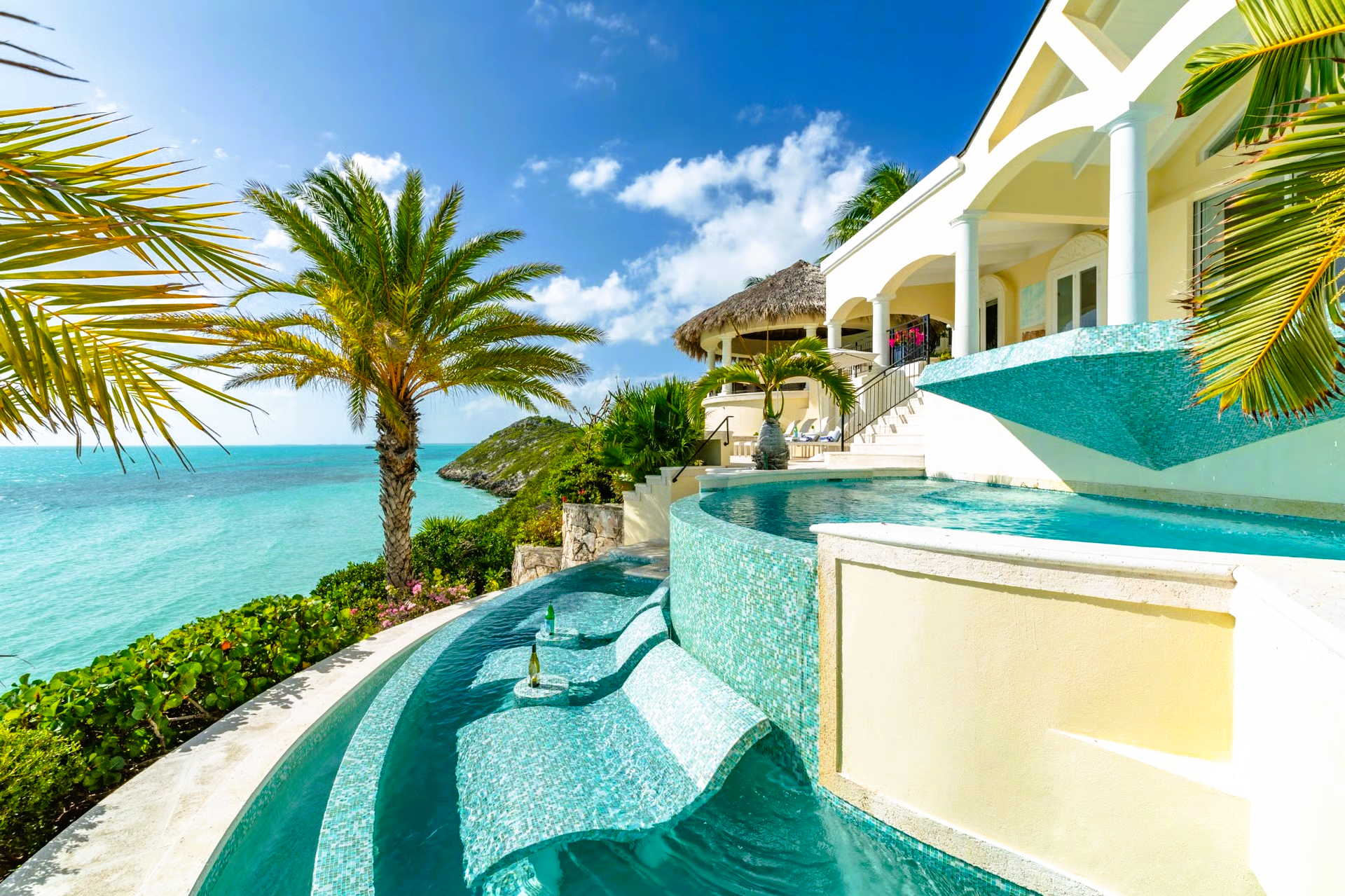 Luxury villa with tiered turquoise pool overlooking ocean, palm trees, and blue sky.