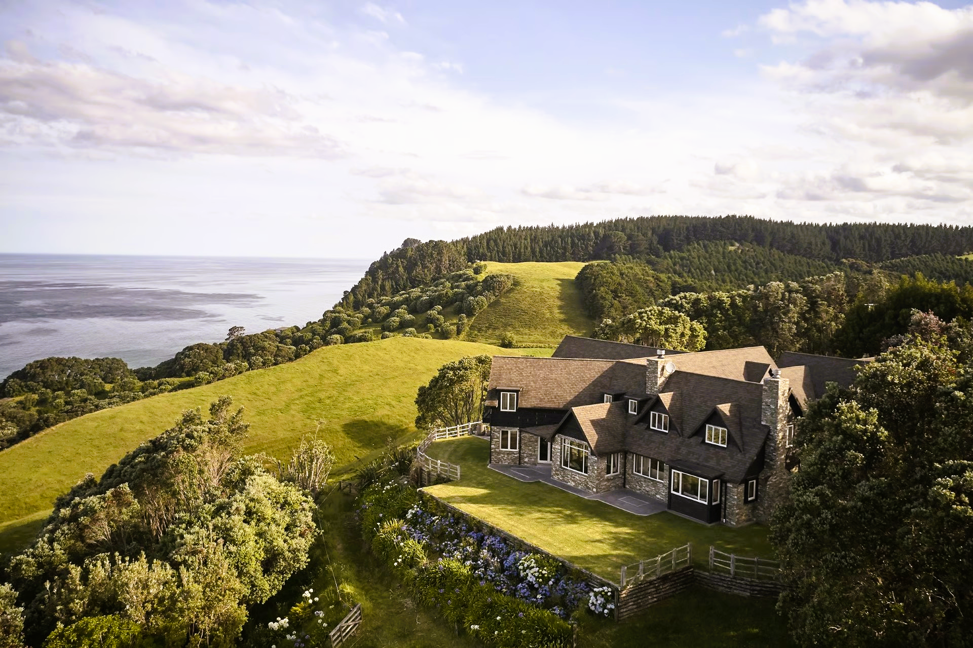 Large stone house on a green hillside overlooking a calm body of water, with forest in the background.