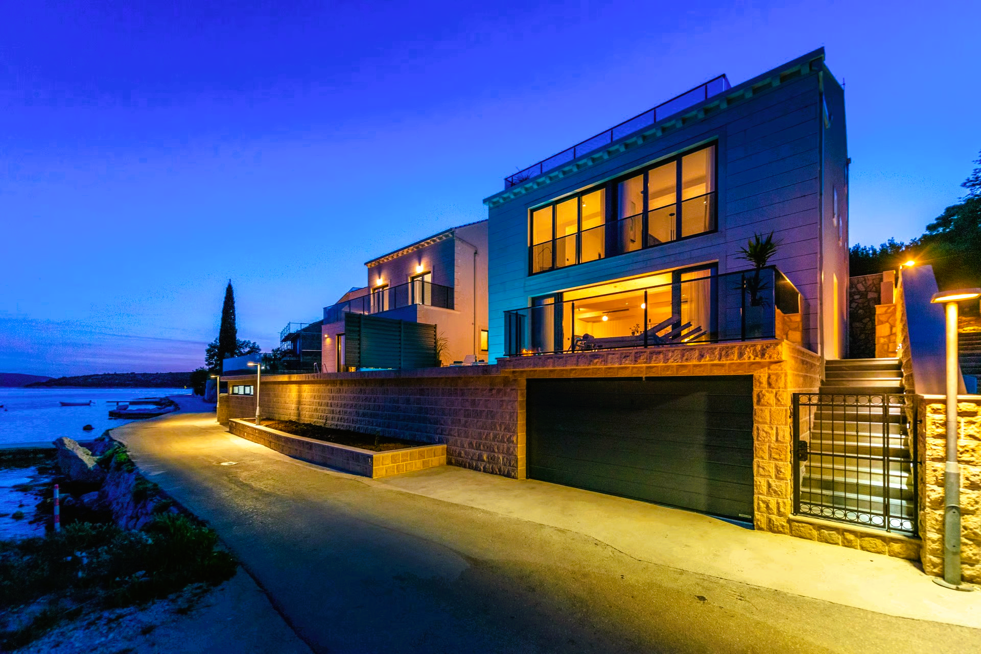 Modern house at dusk with illuminated windows, stone facade, driveway, and waterfront view.