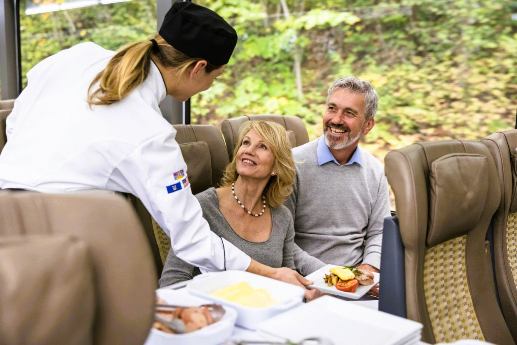 A chef serves a meal to a smiling couple seated inside a train car with a window view.