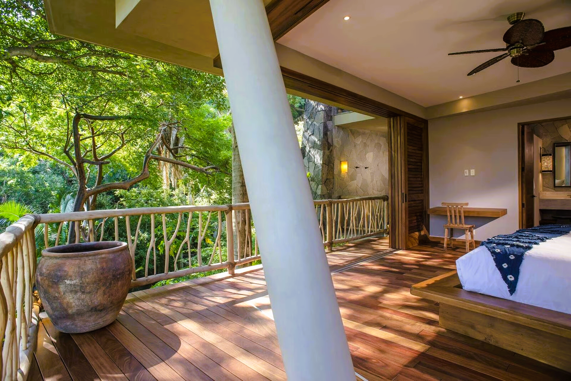 Bedroom with balcony overlooking lush greenery; wooden floor, railing, and furniture.