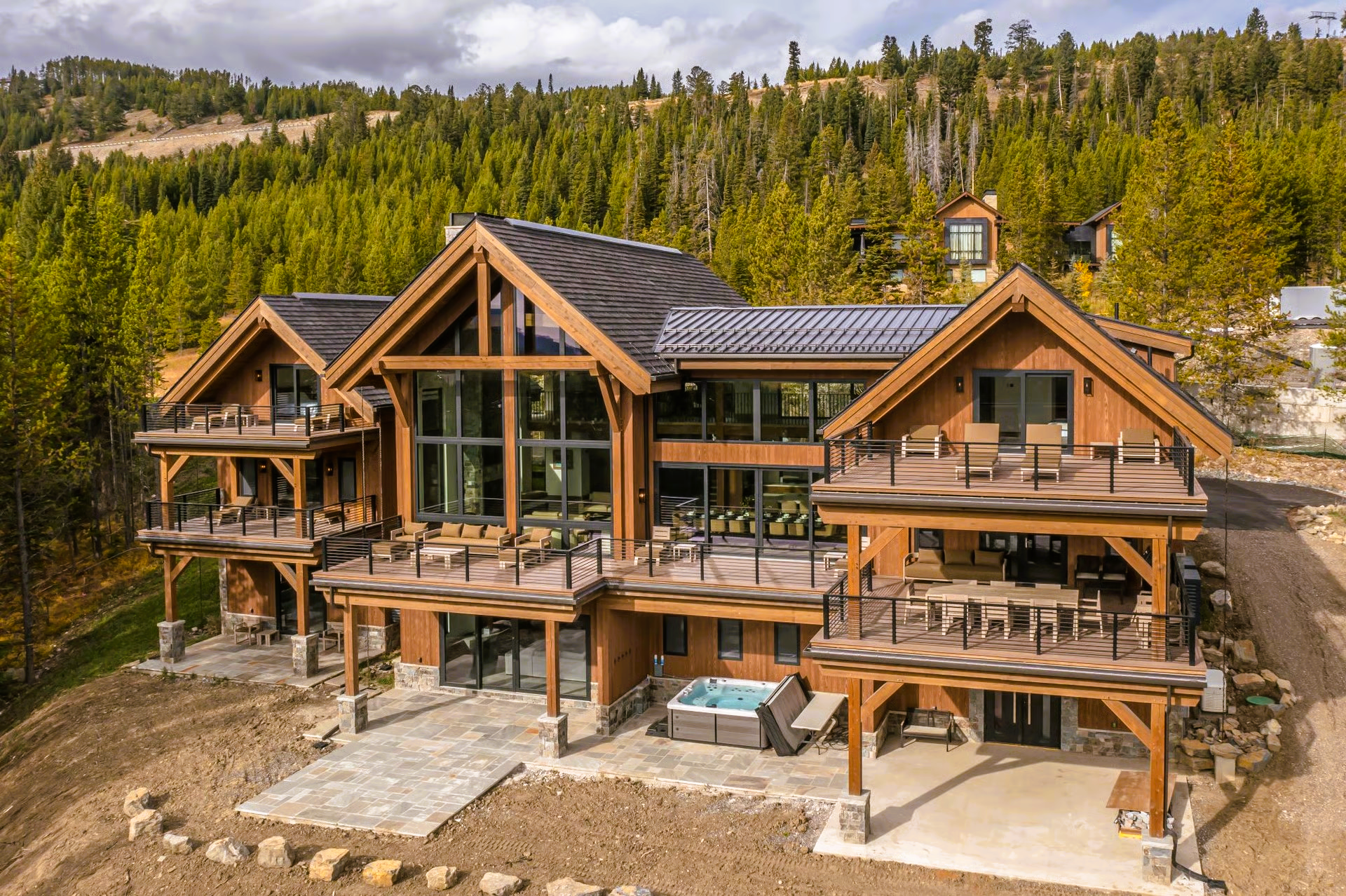 Large brown cabin with multiple decks, hot tub, and a mountain backdrop.