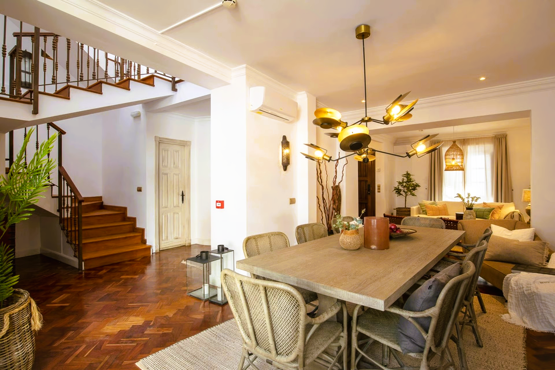 Dining room with wooden table, chairs, chandelier, stairs, and plants.