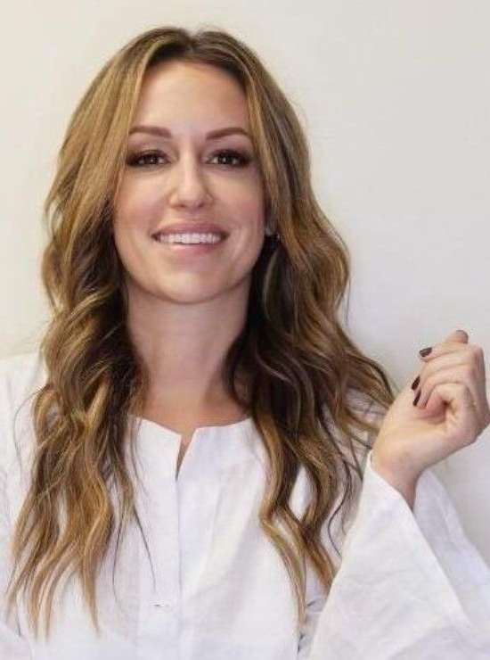 Smiling woman with long wavy hair wearing a white top, against a plain background.