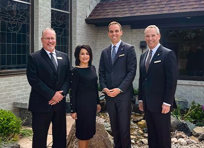 Four formally dressed people pose outdoors near a building and small water feature.
