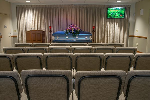 Funeral home chapel with rows of chairs facing a closed blue casket draped with flowers, behind a draped stage.