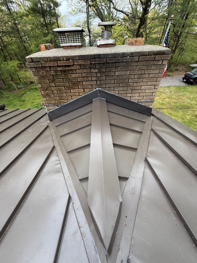 Metal roof peak in foreground, with a brick chimney and stone cap above on a rainy day.