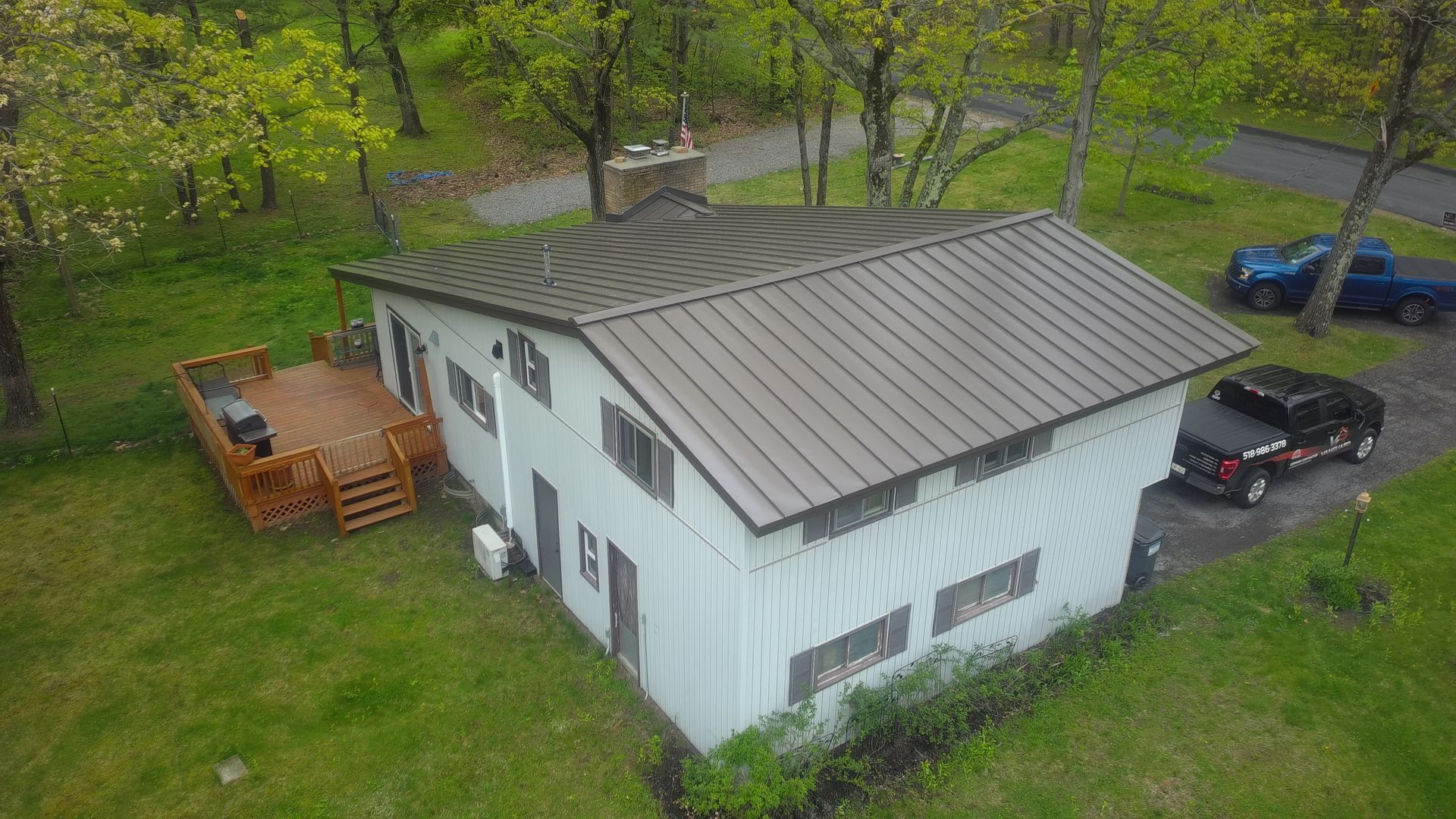 White house with dark roof in a grassy yard, seen from above, with a black car and blue truck nearby.