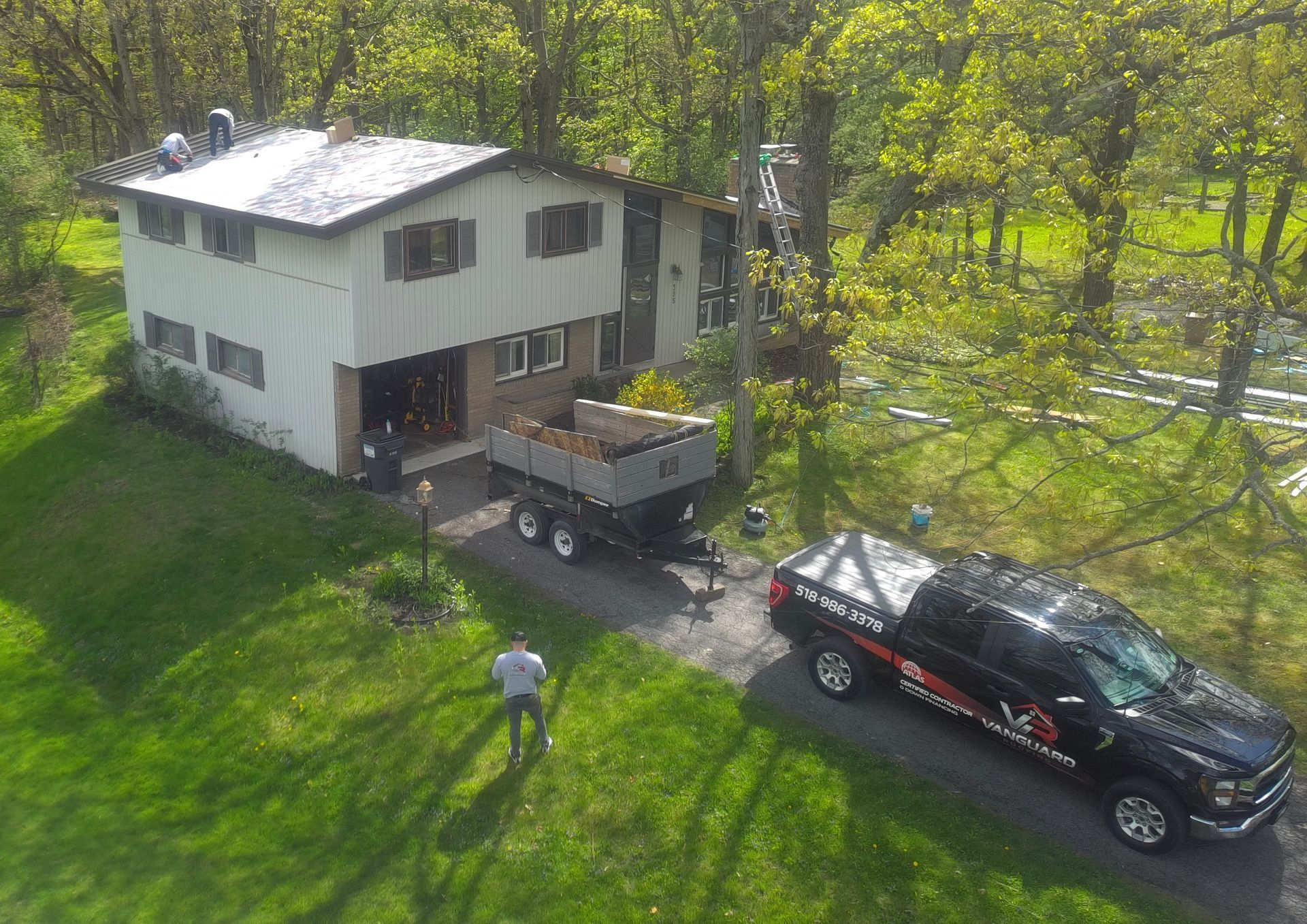 Two-story house with a dump trailer and black truck in a grassy yard; person standing nearby.