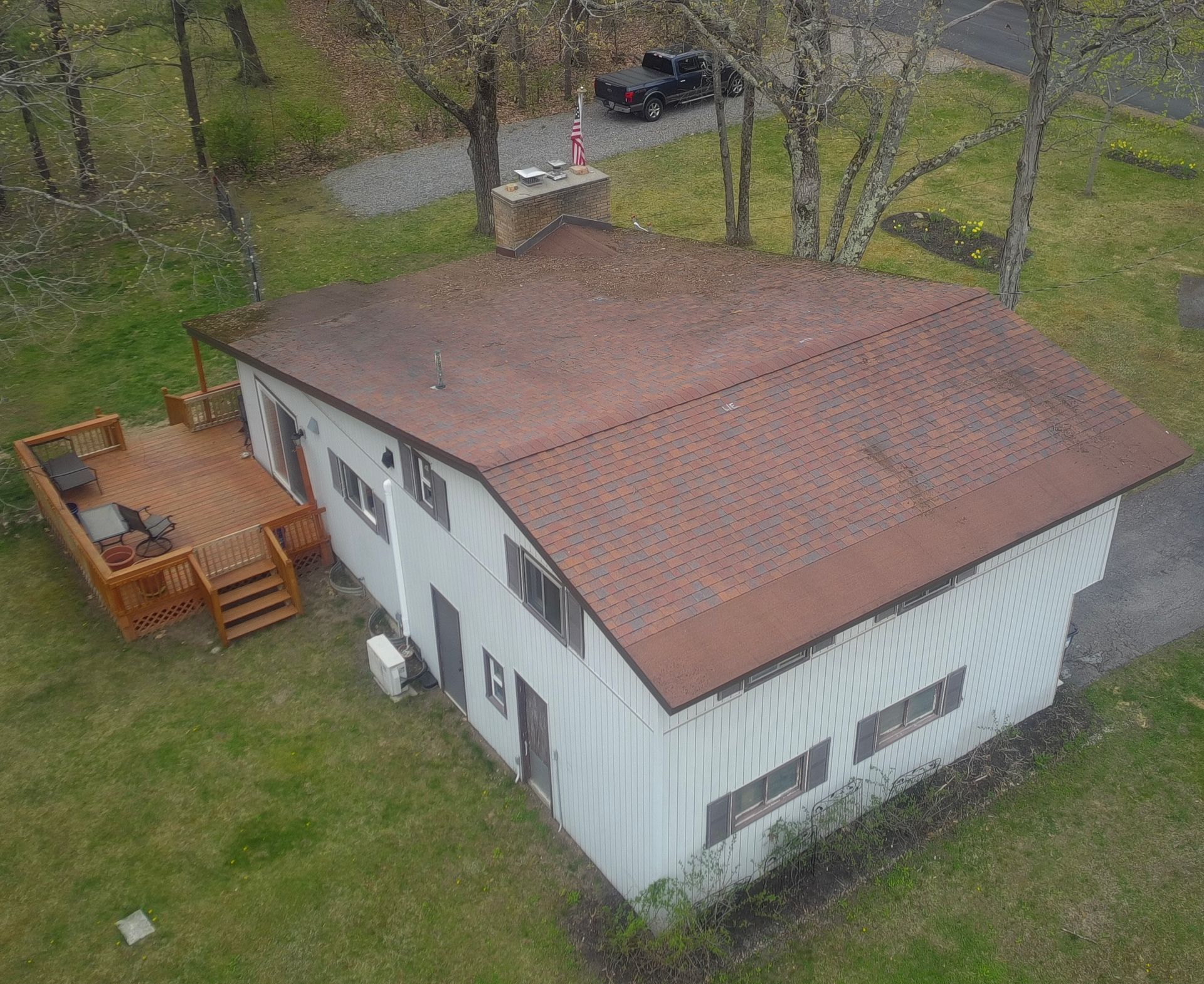 Aerial view of a white house with a brown roof, wooden deck, and grassy yard surrounded by trees.