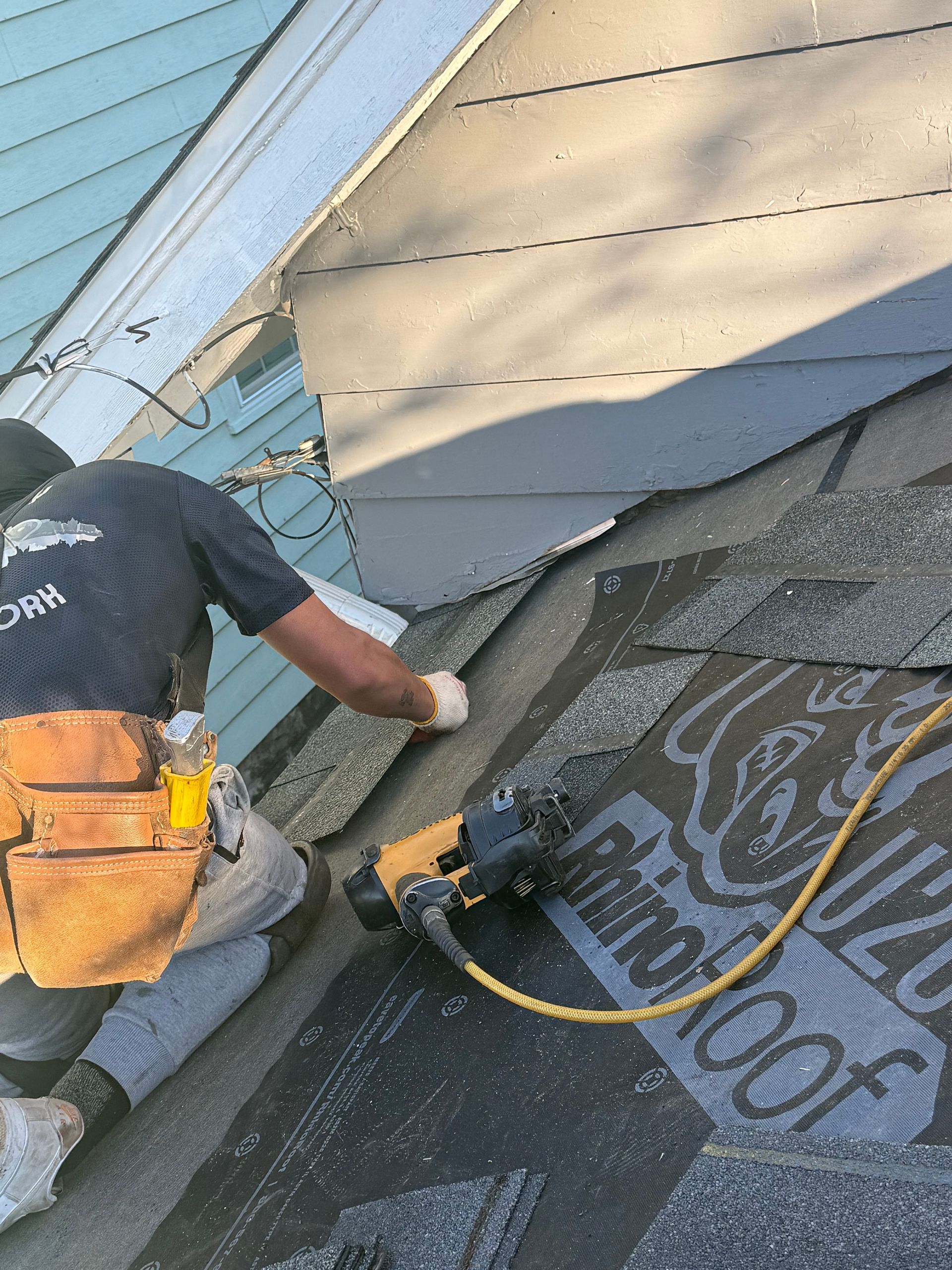 Worker installing roofing underlayment on a sloped roof with a nail gun and yellow hose