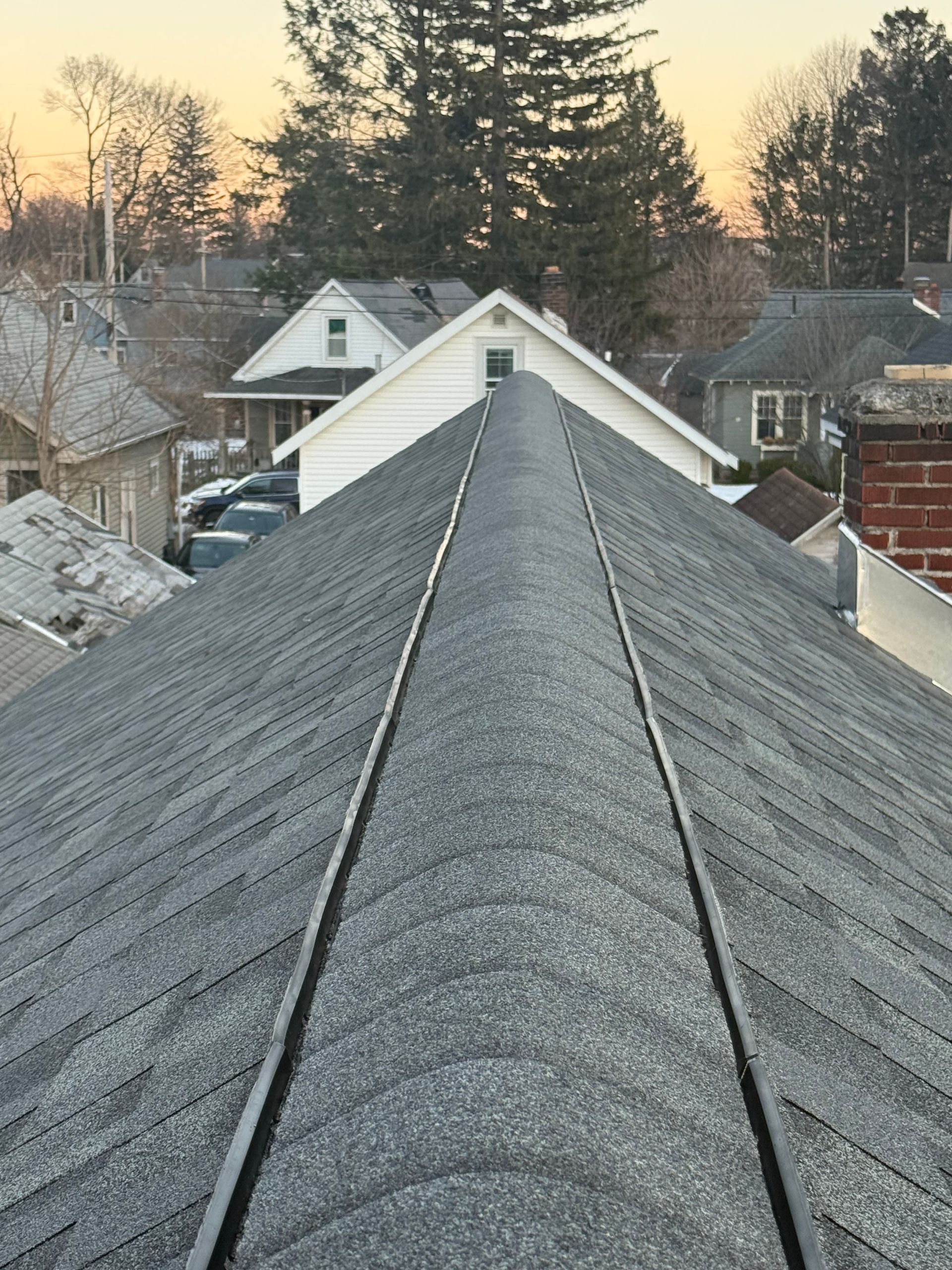 View down a gray shingled roof ridge toward houses and trees at sunset