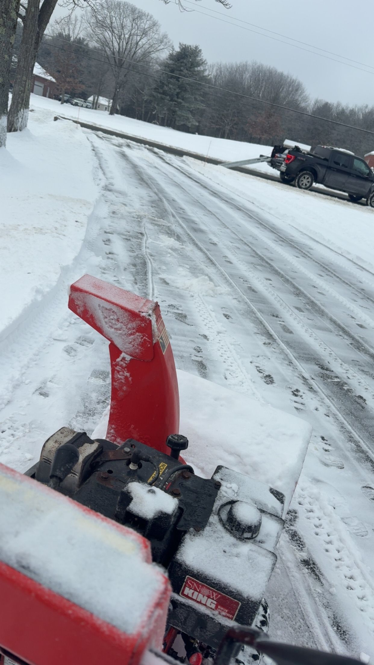 Snow blower clearing a snow-covered roadside with cars parked in the distance.