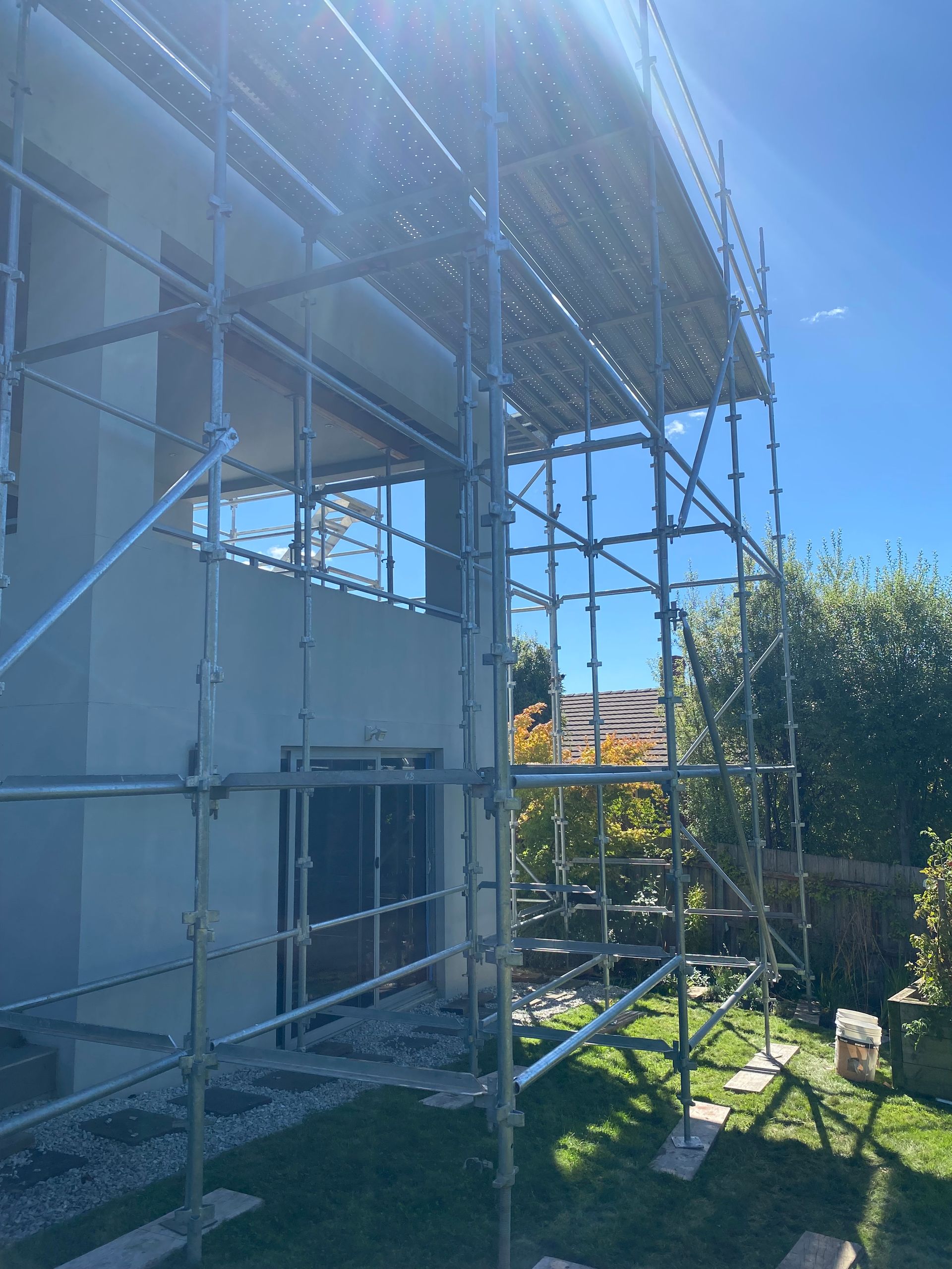Scaffolding beside a house wall in bright sunlight, with grass and trees nearby