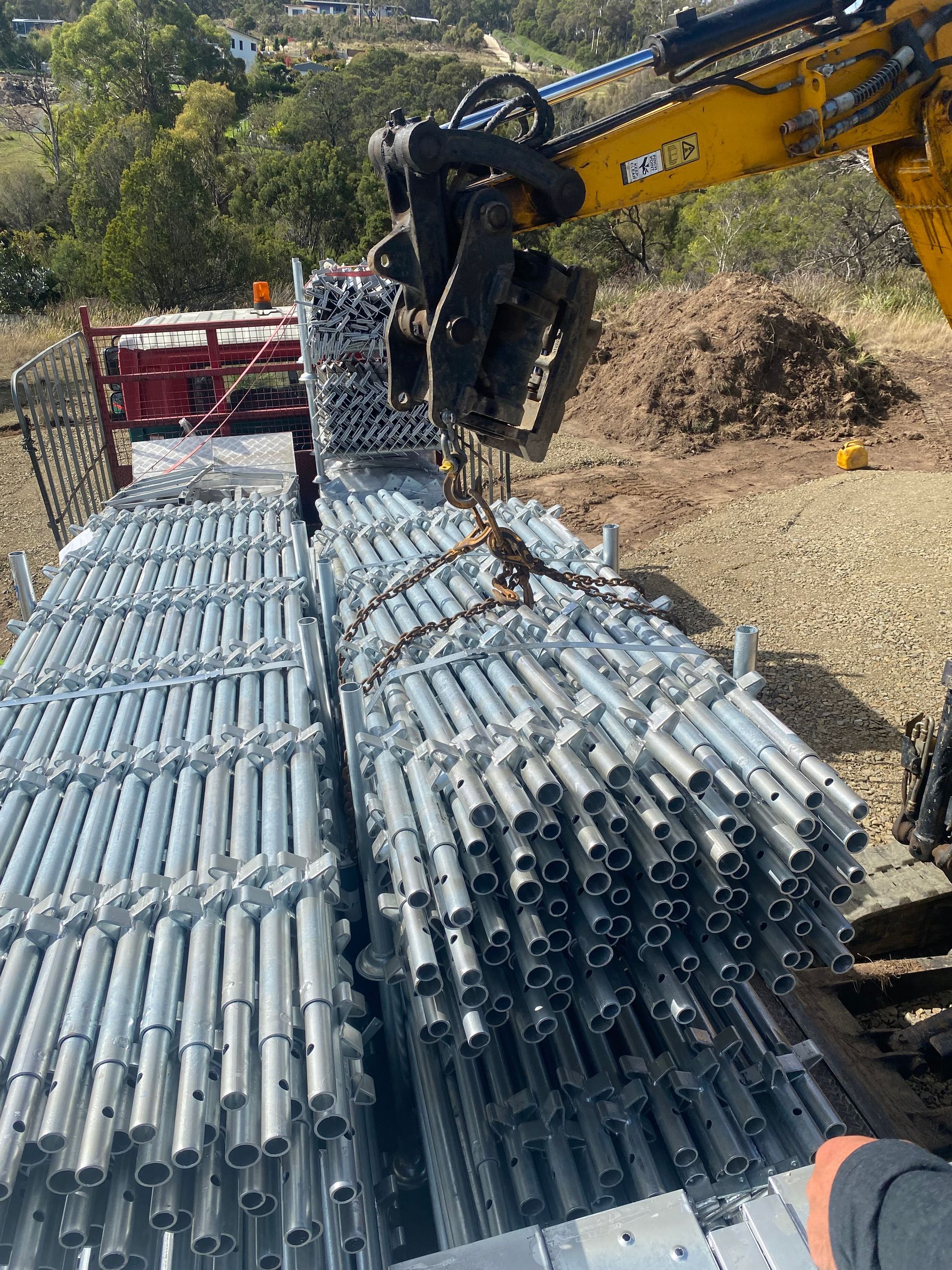 Excavator claw lifting stacked metal scaffolding pipes outdoors on a gravel site