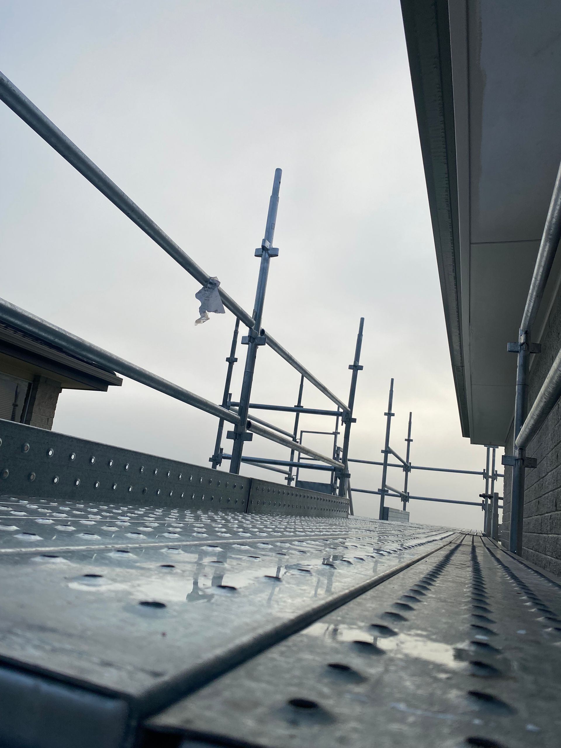 Metal rooftop walkway with scaffolding and wet riveted floor under an overcast sky