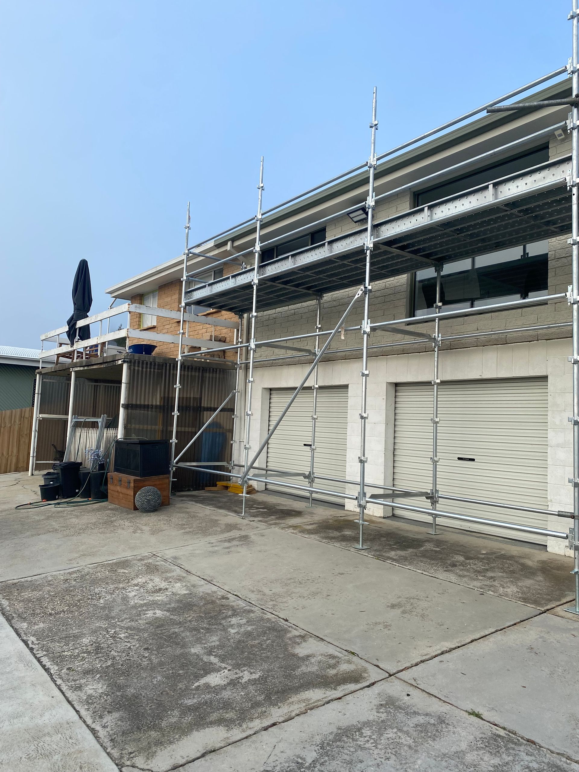 Scaffolding along a building exterior with boarded windows and a concrete driveway under a blue sky