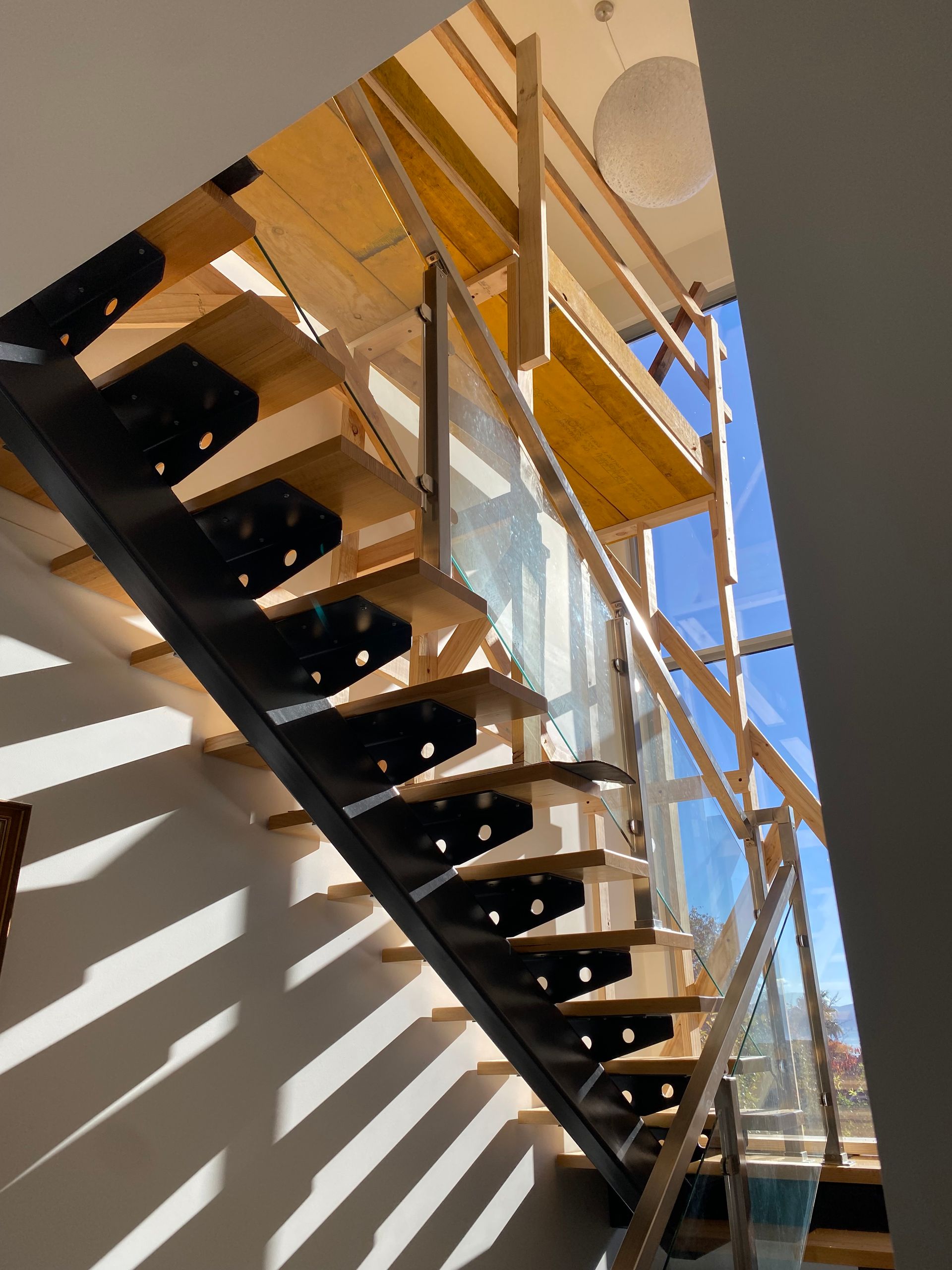 Modern staircase with black treads, white wall, glass railings, and sunlit shadows against a blue sky.