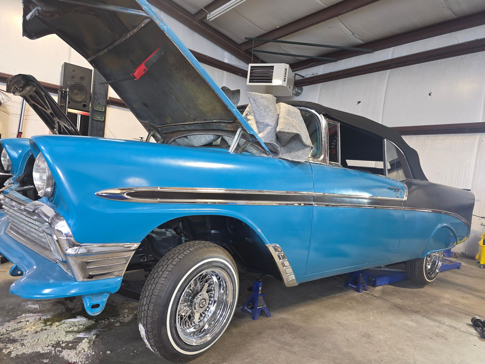 Blue classic car with hood open in a garage, showing damage and missing wheels.