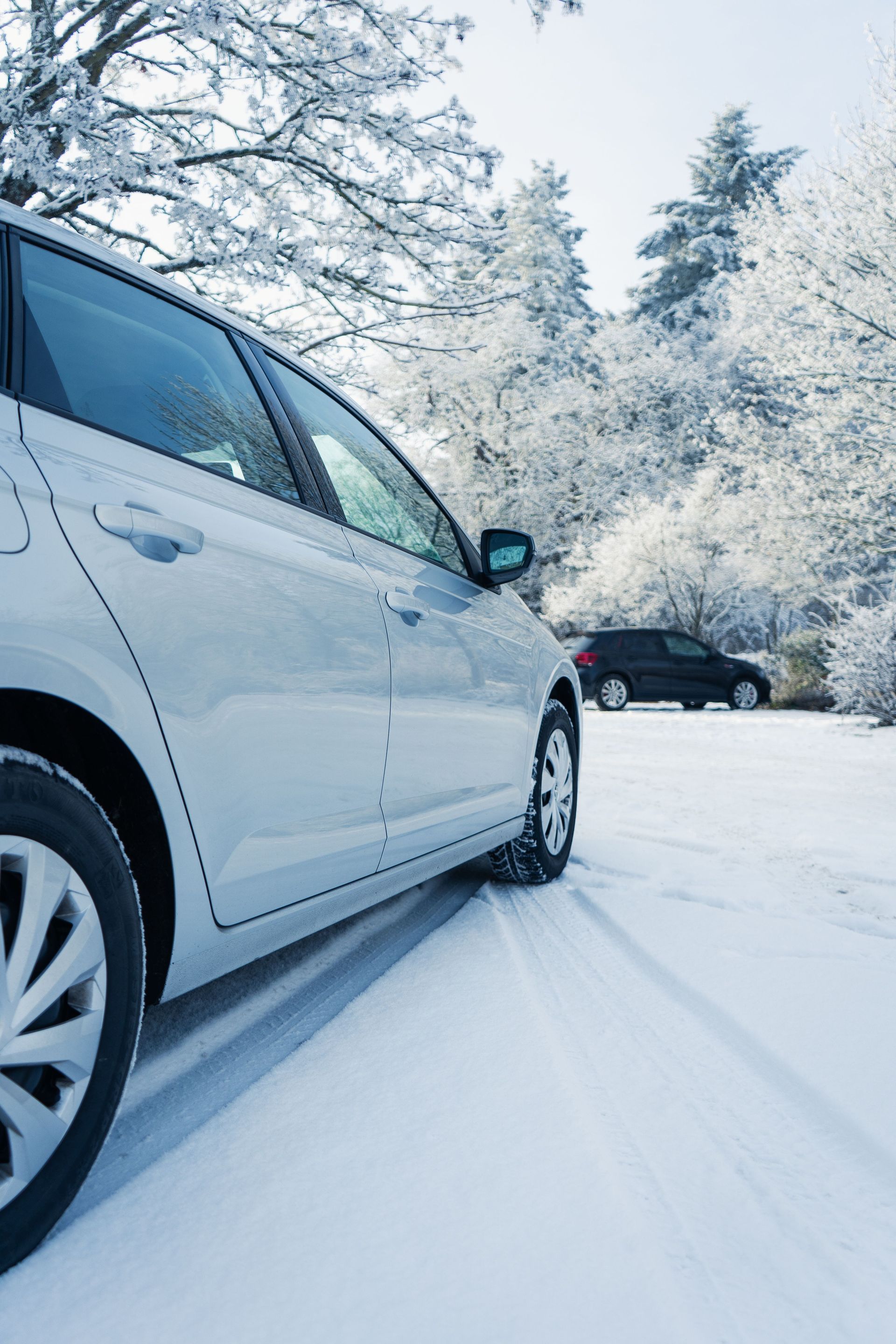 White car parked on a snowy road beside frosted trees, with a dark car in the background