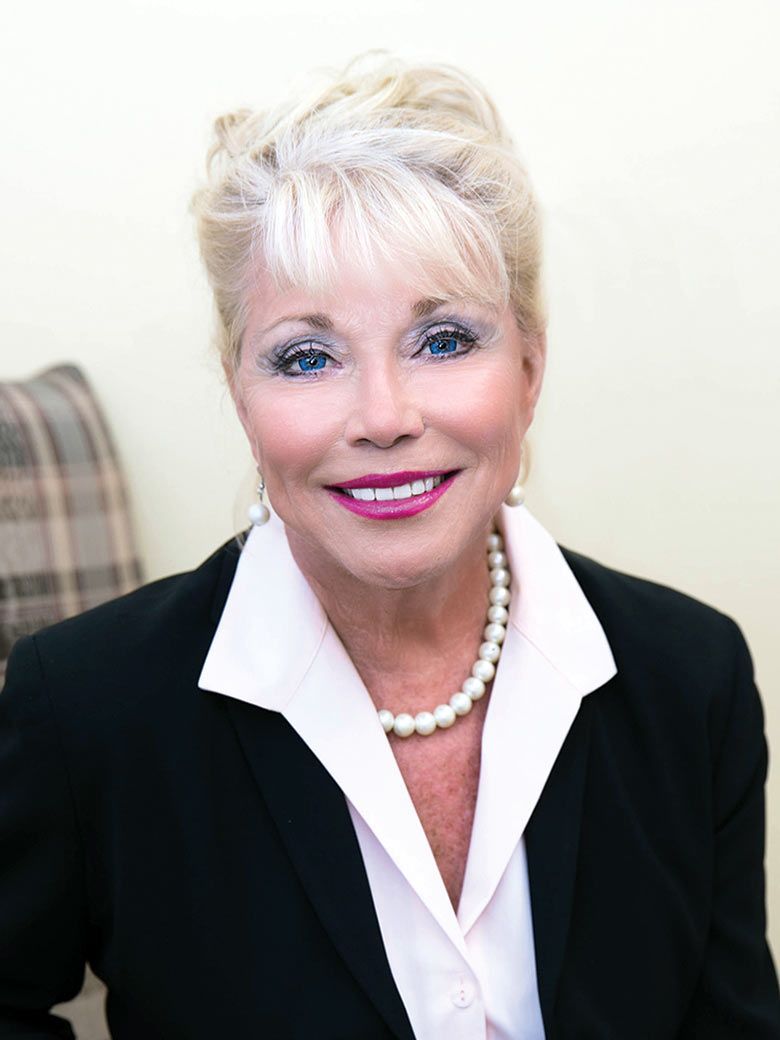 A woman wearing a black jacket and pearls smiles for the camera