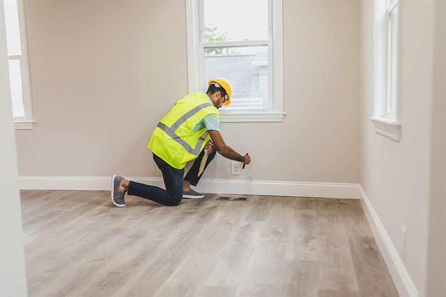 A person in a yellow hard hat and safety vest kneels on a light wood floor, inspecting an air vent near the baseboard.