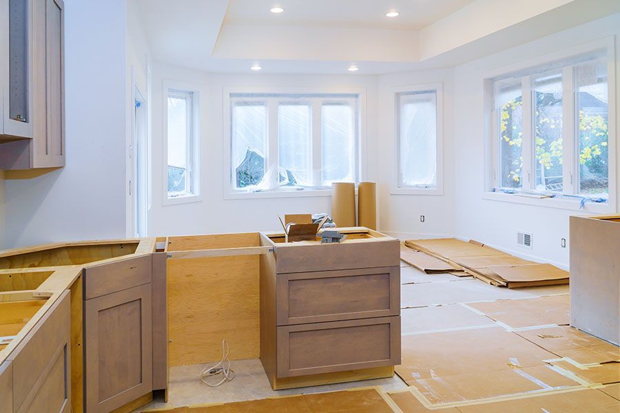An unfinished kitchen renovation features light-colored wooden cabinets installed against white walls and covered floors.