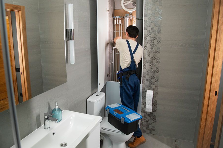 A technician in work coveralls repairs plumbing behind a bathroom wall panel, standing on a closed toilet seat.