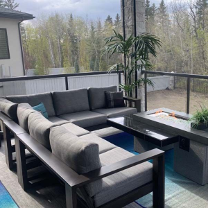 A grey patio sectional and fire pit table set on a balcony overlooking a wooded backyard.