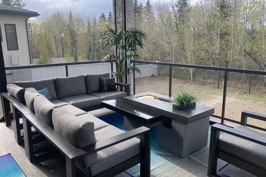 A modern outdoor patio set with gray sectional sofas and a rectangular fire pit table on a deck overlooking a wooded yard.