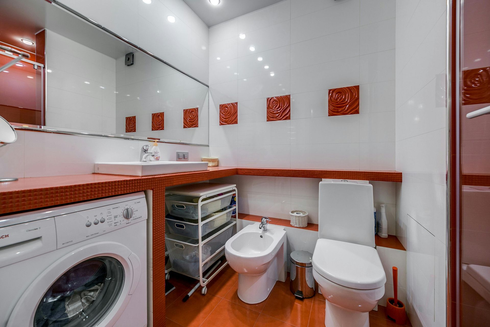 A modern bathroom featuring a white toilet, bidet, and washing machine beneath a wooden vanity counter with wall decor.