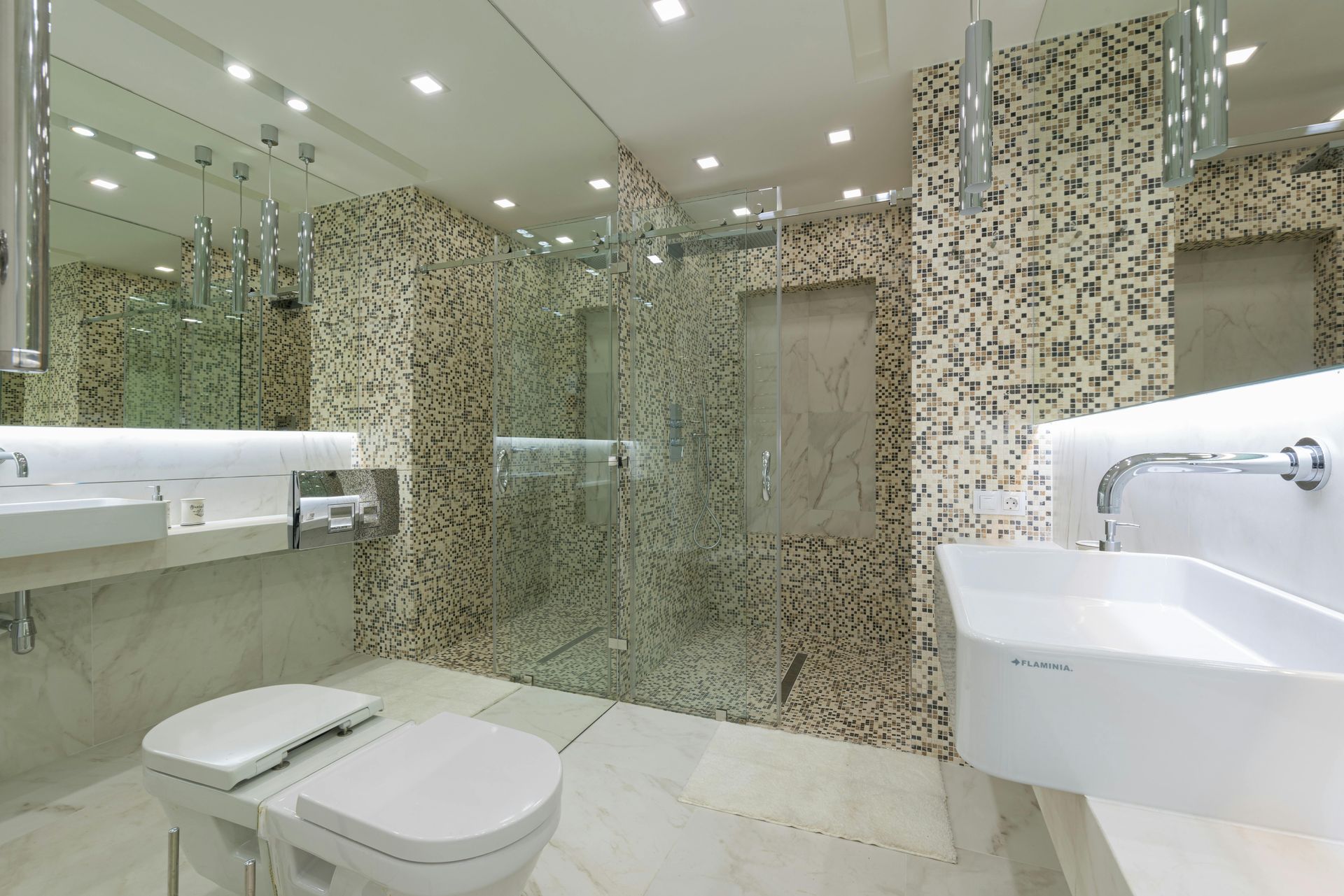 A modern bathroom featuring two white pedestal sinks, a toilet, and a glass-enclosed shower with decorative stone-tile walls.